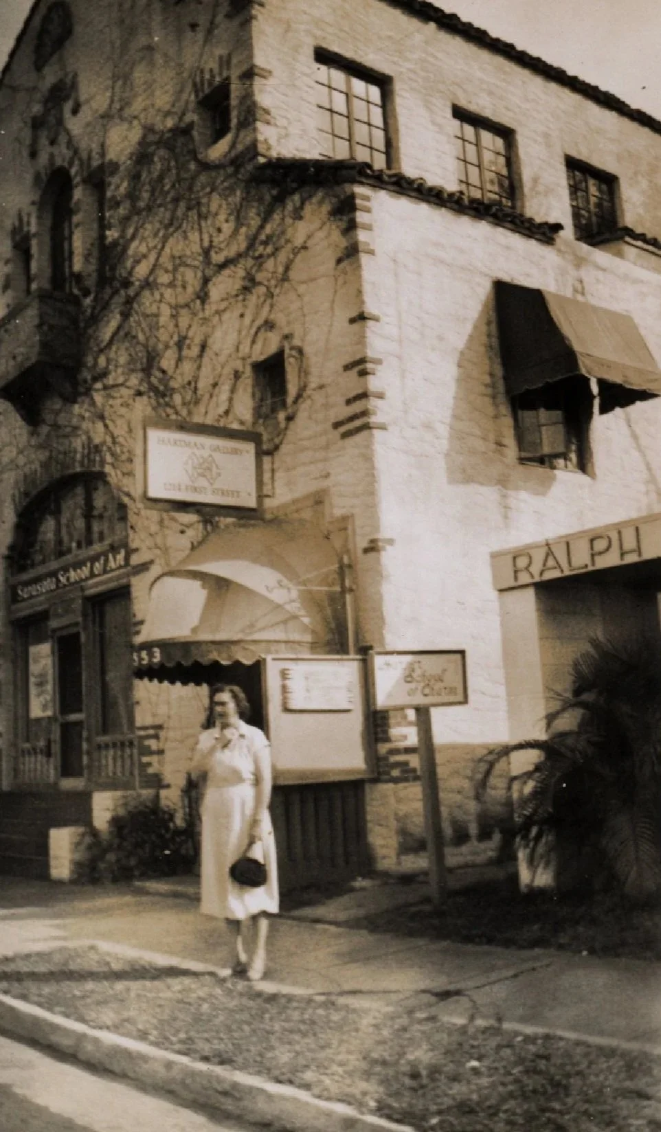 A woman standing outside a building with a sign that reads 'Ralph.' The building has a sign pointing to Hartman Gallery on First Street and another sign for School of Charm. The woman is wearing a light-colored dress and holding a small purse, and there is a palm plant in front of the building.