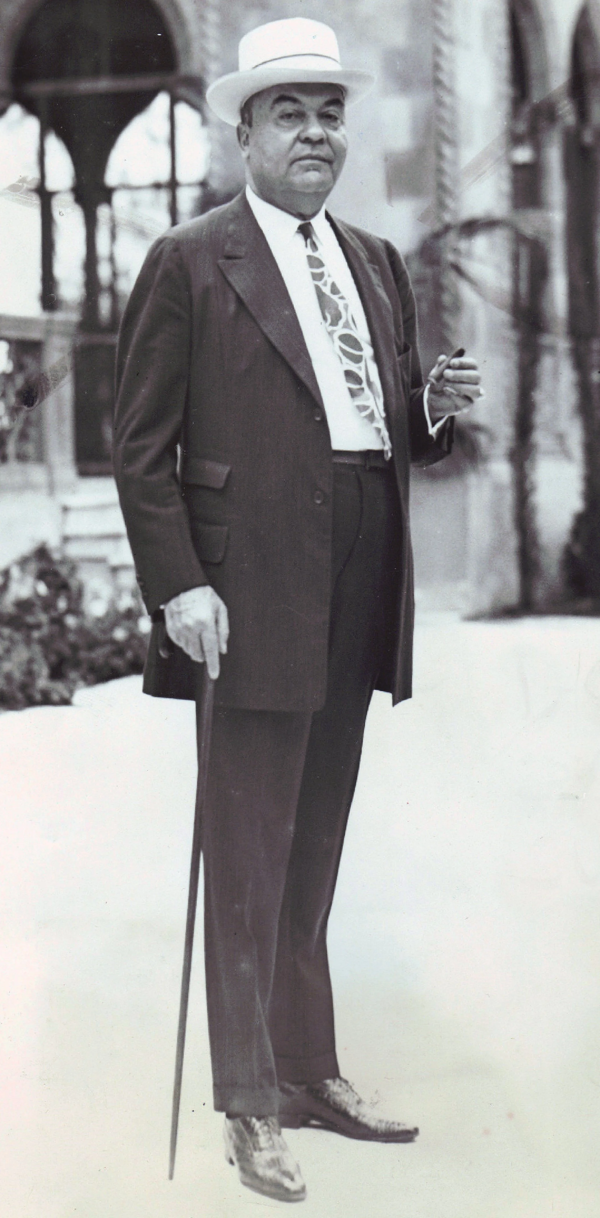 A distinguished man wearing a suit, tie, overcoat, and a fedora hat, standing outdoors with a cane in one hand and a cigar in the other, in front of a building with arched windows.