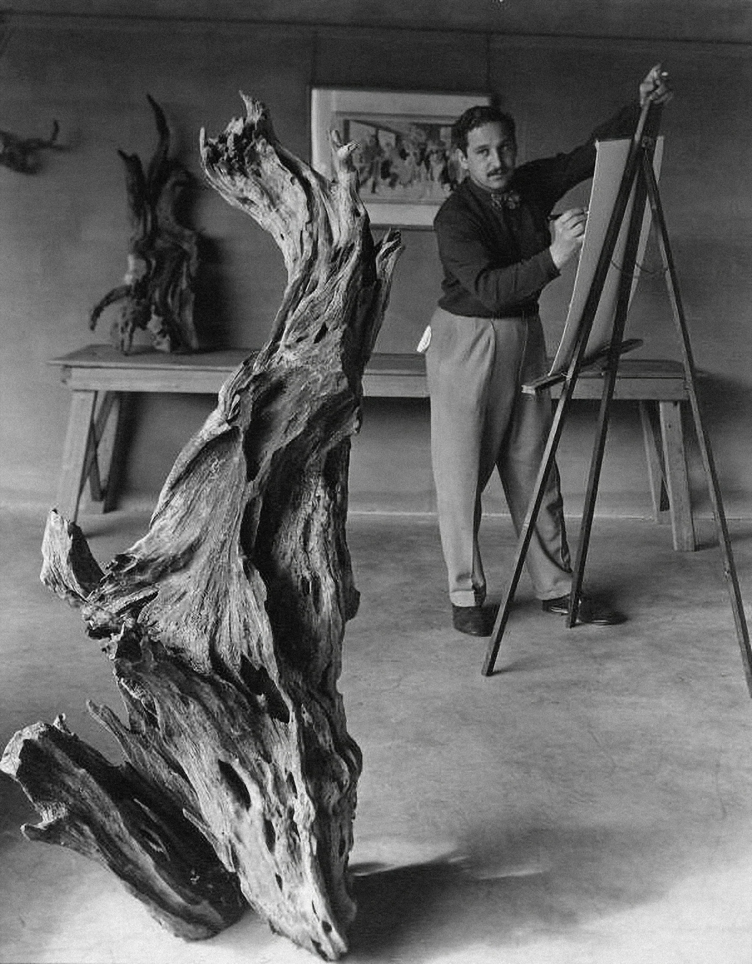 A man with a mustache and dark hair drawing on an easel in a room with wood-paneled walls, with a large, twisted piece of driftwood or decorative wood sculptural art in the foreground and a framed picture on the wall behind him.