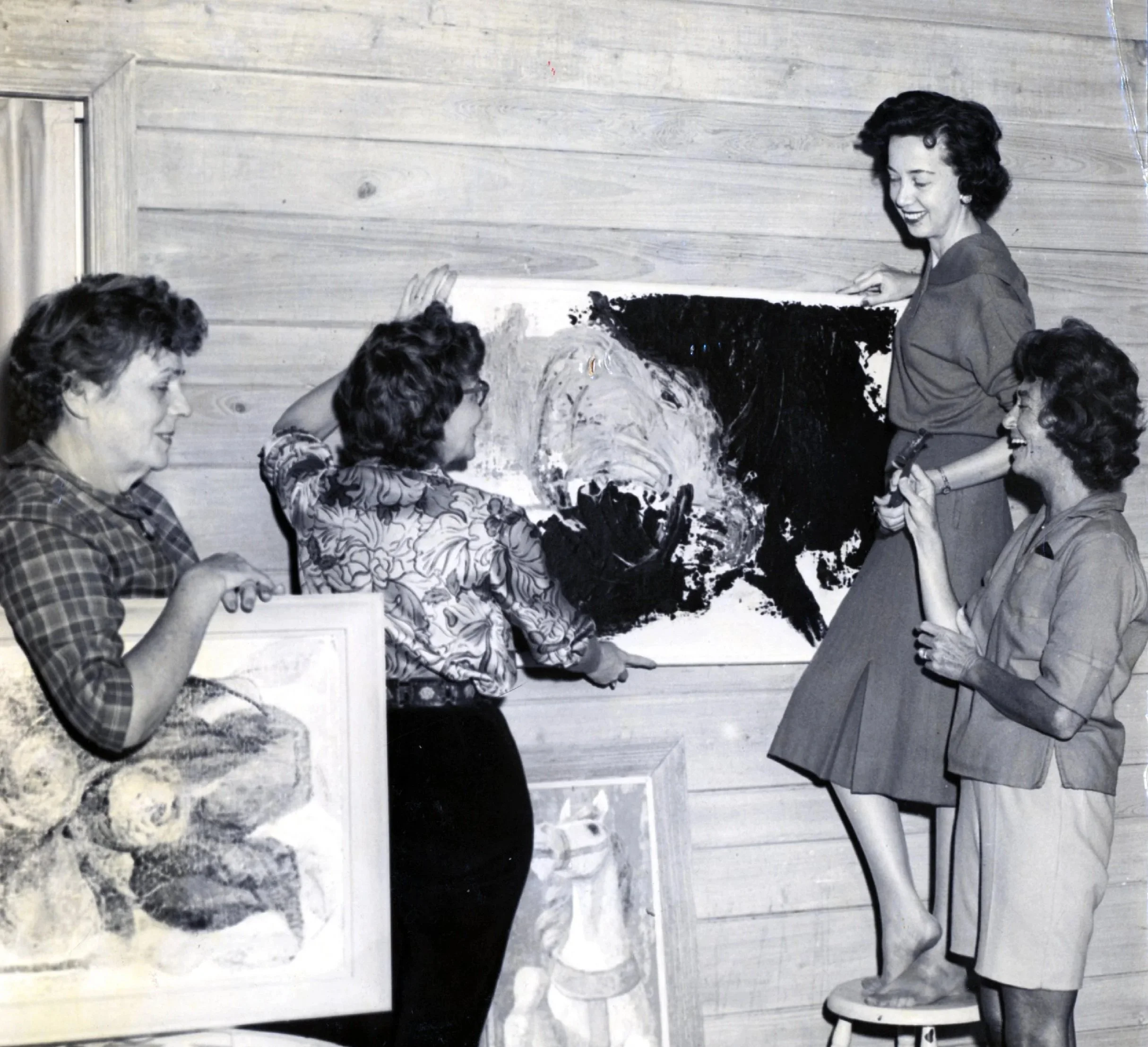 Group of women viewing and discussing paintings in an art gallery, with one woman standing on a stool to examine a large abstract black and white artwork.