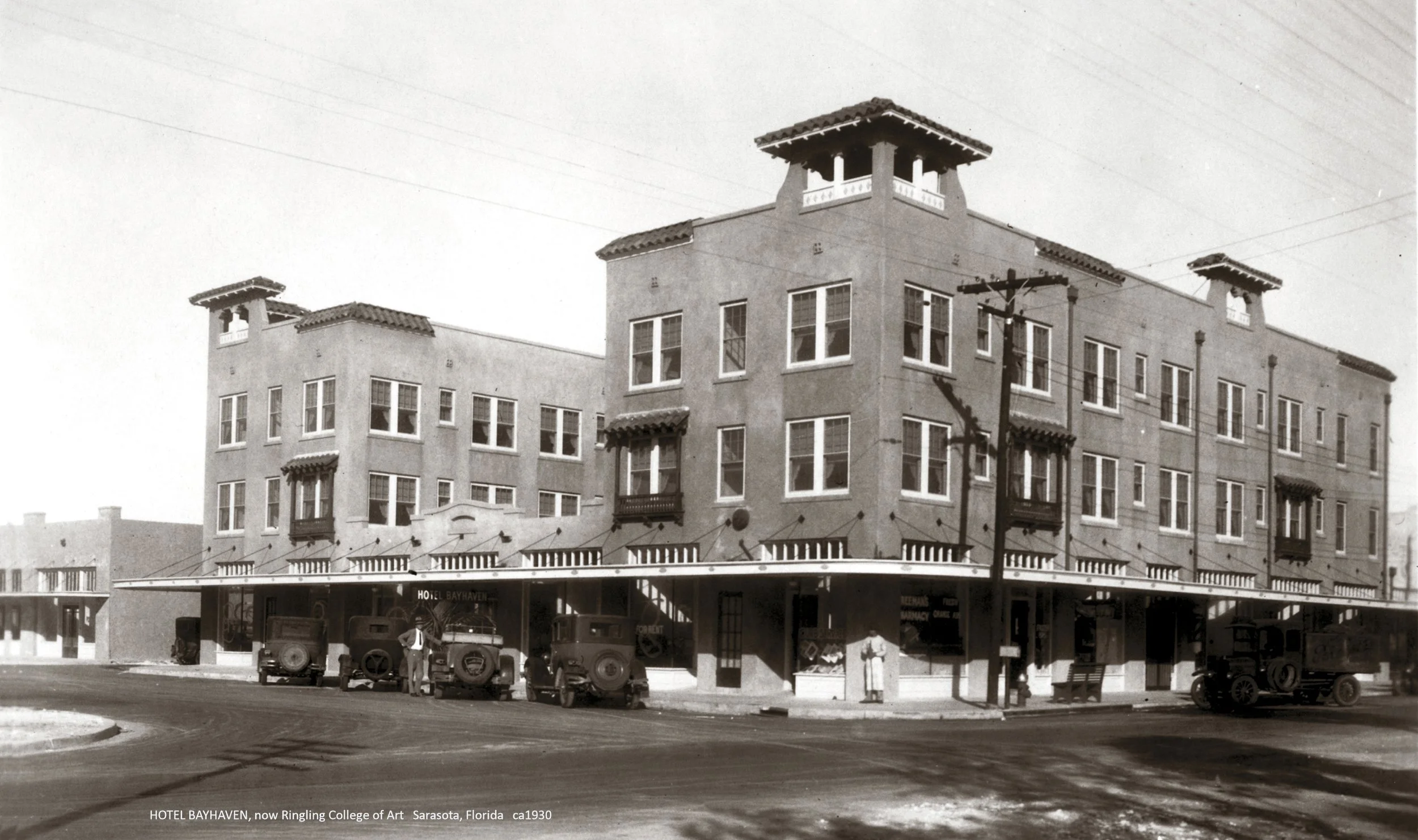 Black and white photo of HOTEL BAYHAVEN, now Ringling College of Art in Sarasota, Florida, circa 1930. The building is a large, multi-story structure with a flat roof and small decorative towers at the corners. Several vintage cars are parked in front, and a few people are visible near the entrance, with a streetlamp and overhead utility lines also in view.