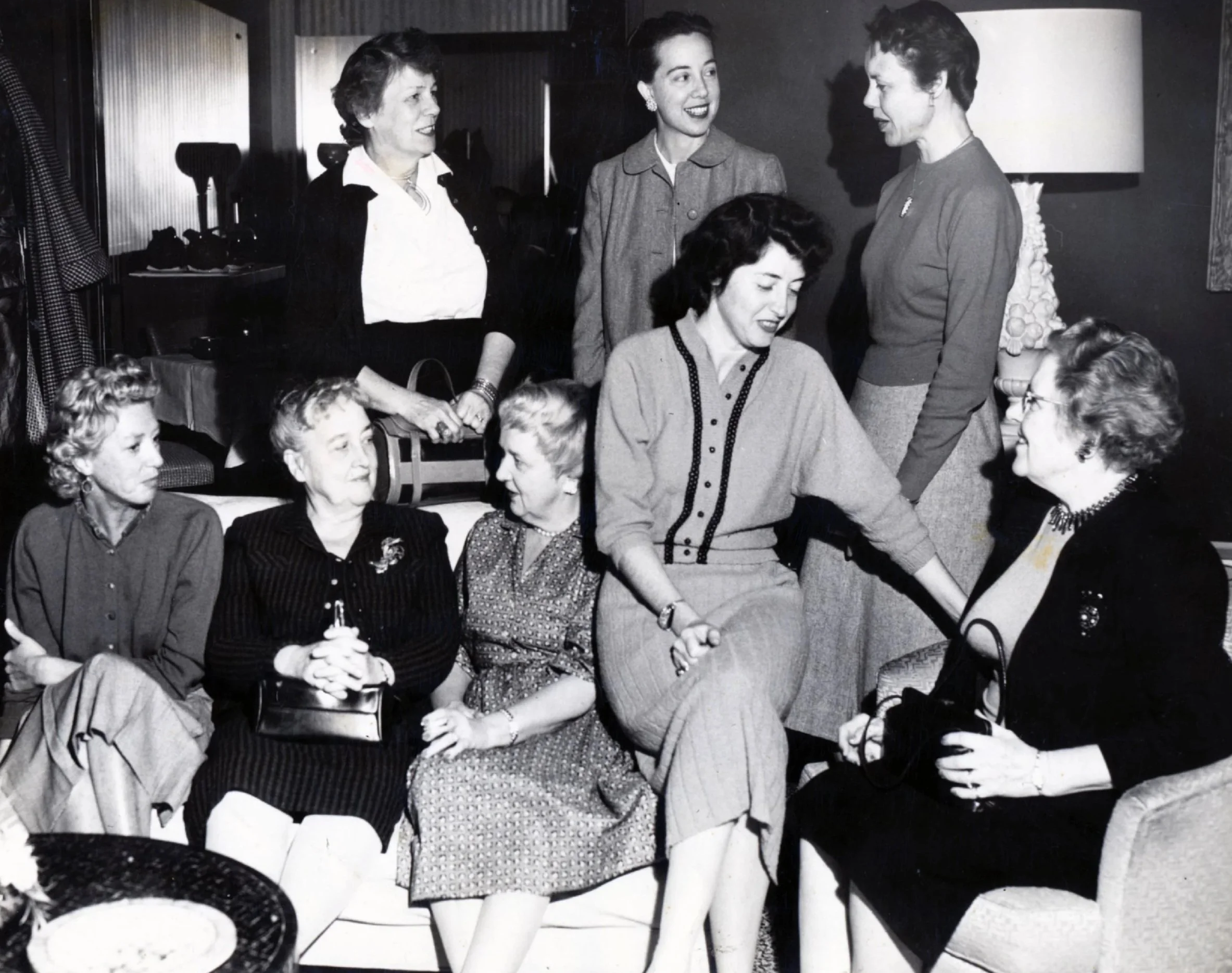 Vintage black-and-white photo of eight women, some sitting on a sofa and others standing behind, engaged in conversation in a living room setting with a lamp and a table.