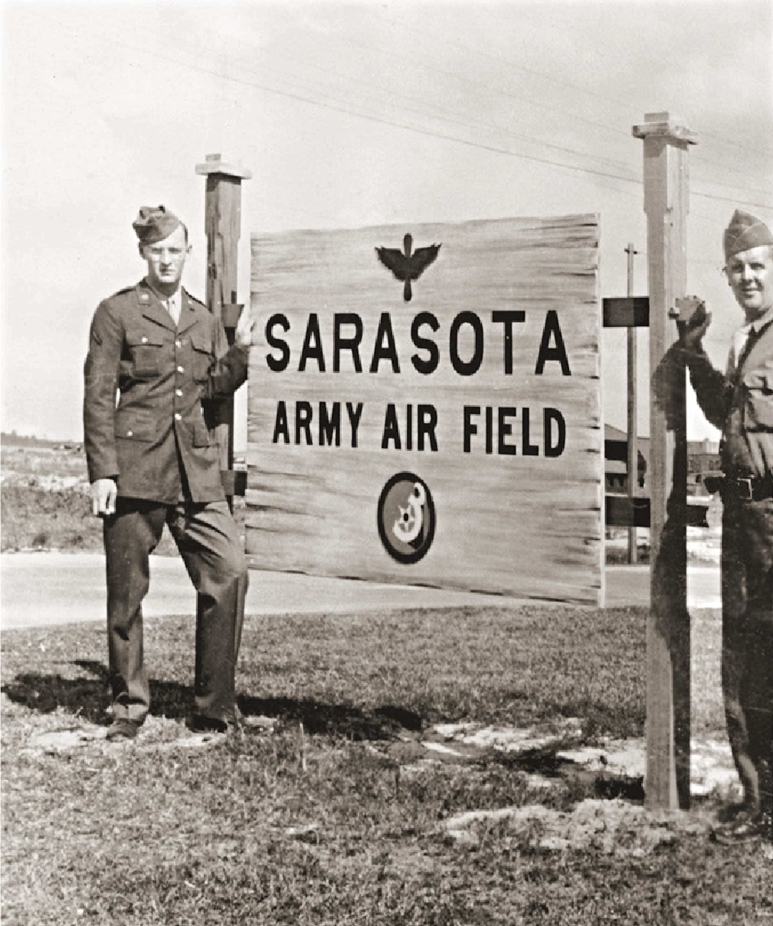 Two soldiers in uniform standing next to a large wooden sign that reads 'Sarasota Army Air Field' with symbols of a bird and an aircraft.