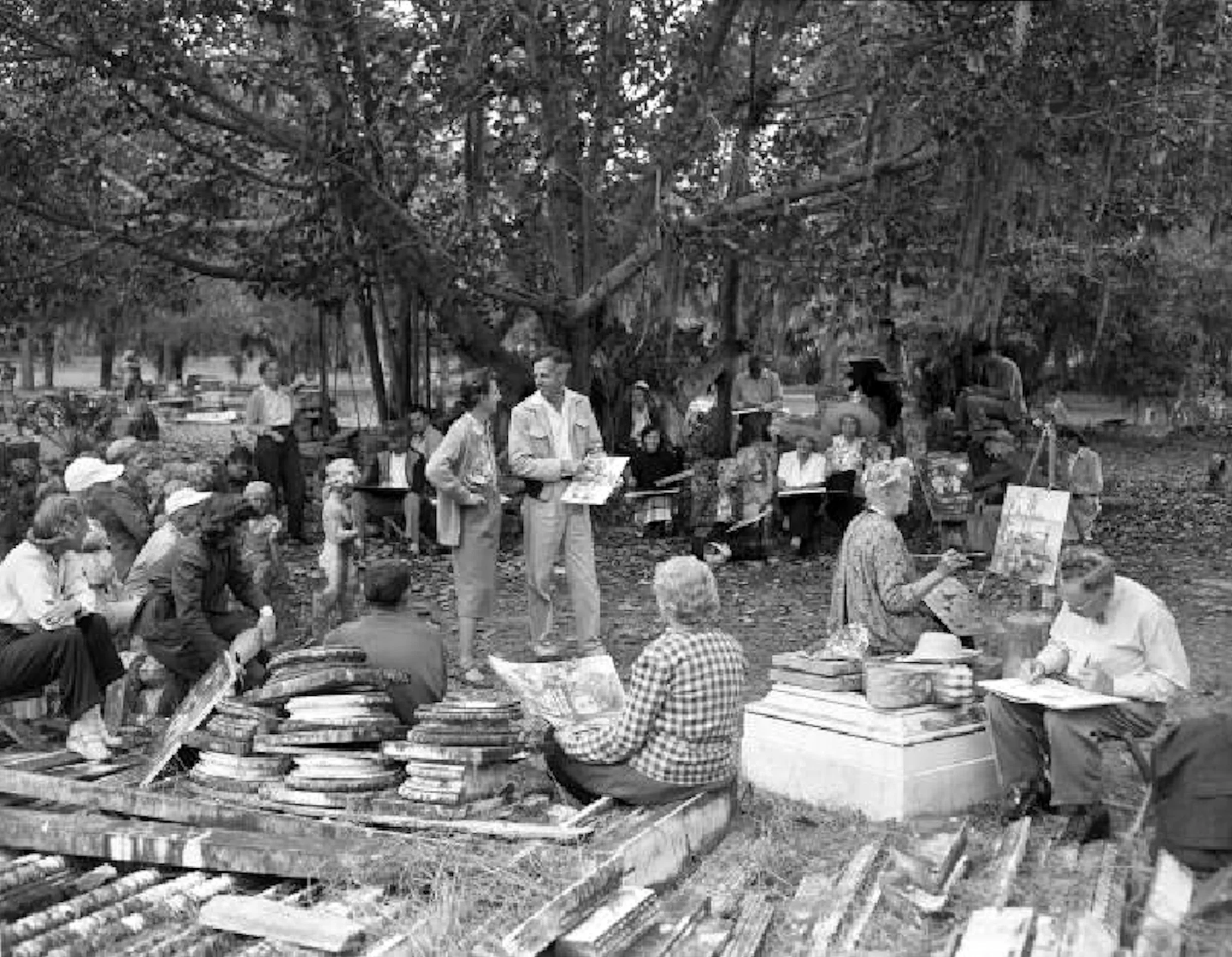 Black and white photo of an outdoor art class or workshop in a park with many people, some drawing or painting at easels, others observing. Large trees in the background provide shade, with supplies and artwork visible.