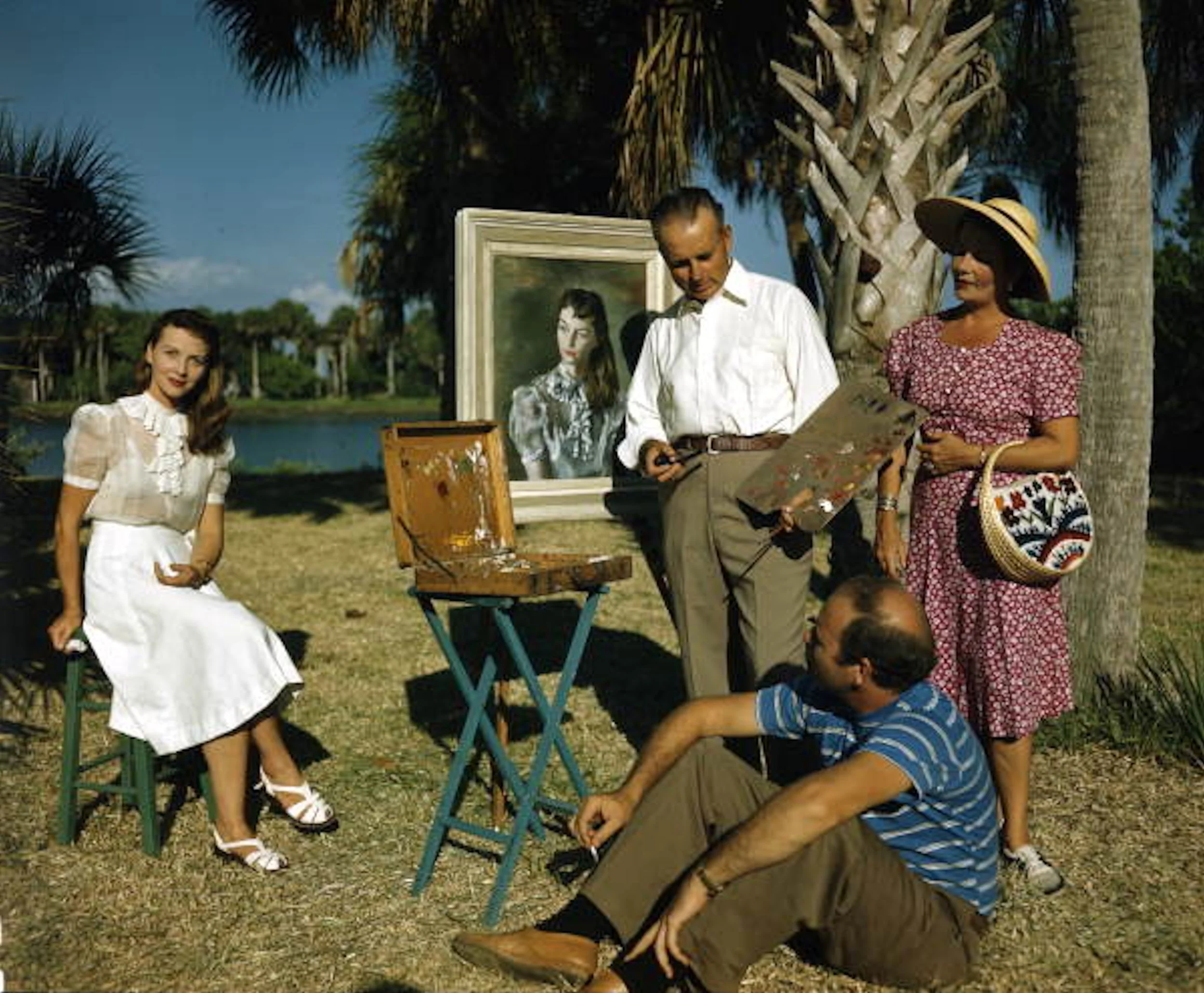 A group of five people outdoors around a painting. A woman in a white dress is sitting on a stool, a man in a white shirt is holding a palette and camera, a woman in a pink dress with a large hat and bag is standing, and a man in a blue striped shirt is sitting on the ground. There are palm trees and a lake in the background.