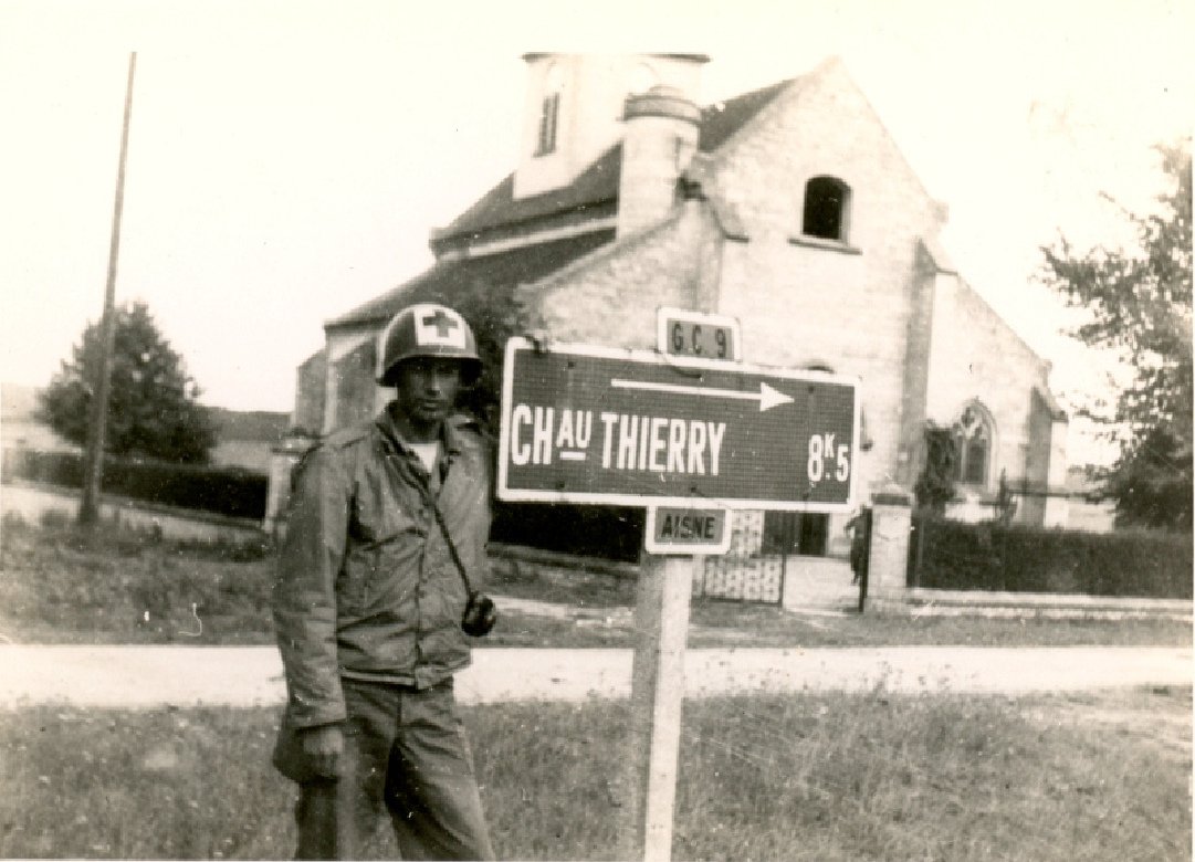 A soldier in uniform and helmet standing near a street sign that says 'Chau Thierry 0.5'. A church and a person in the background.