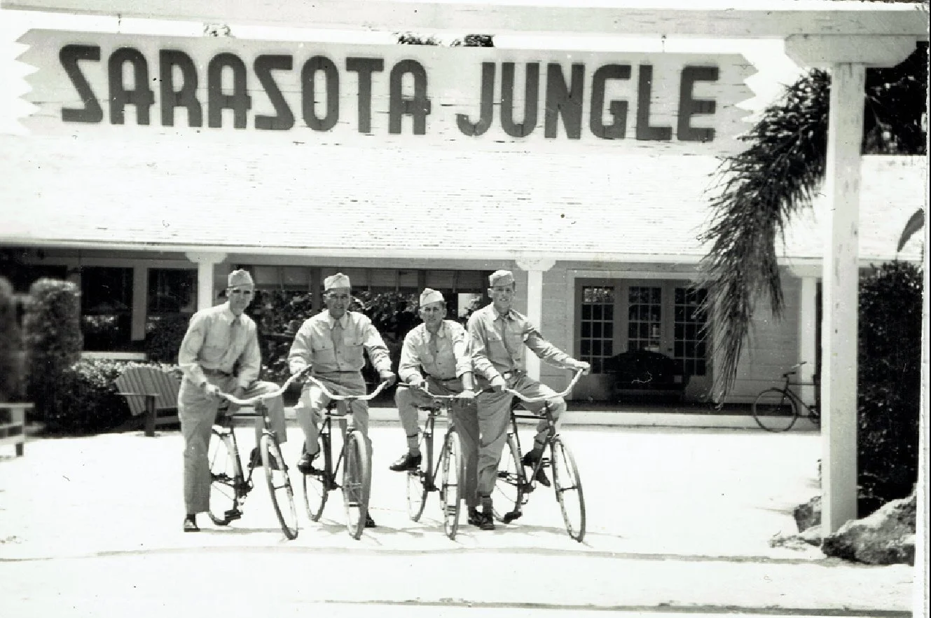 Four men in uniform riding bicycles under a large sign that reads 'Sarasota Jungle' in front of a building.