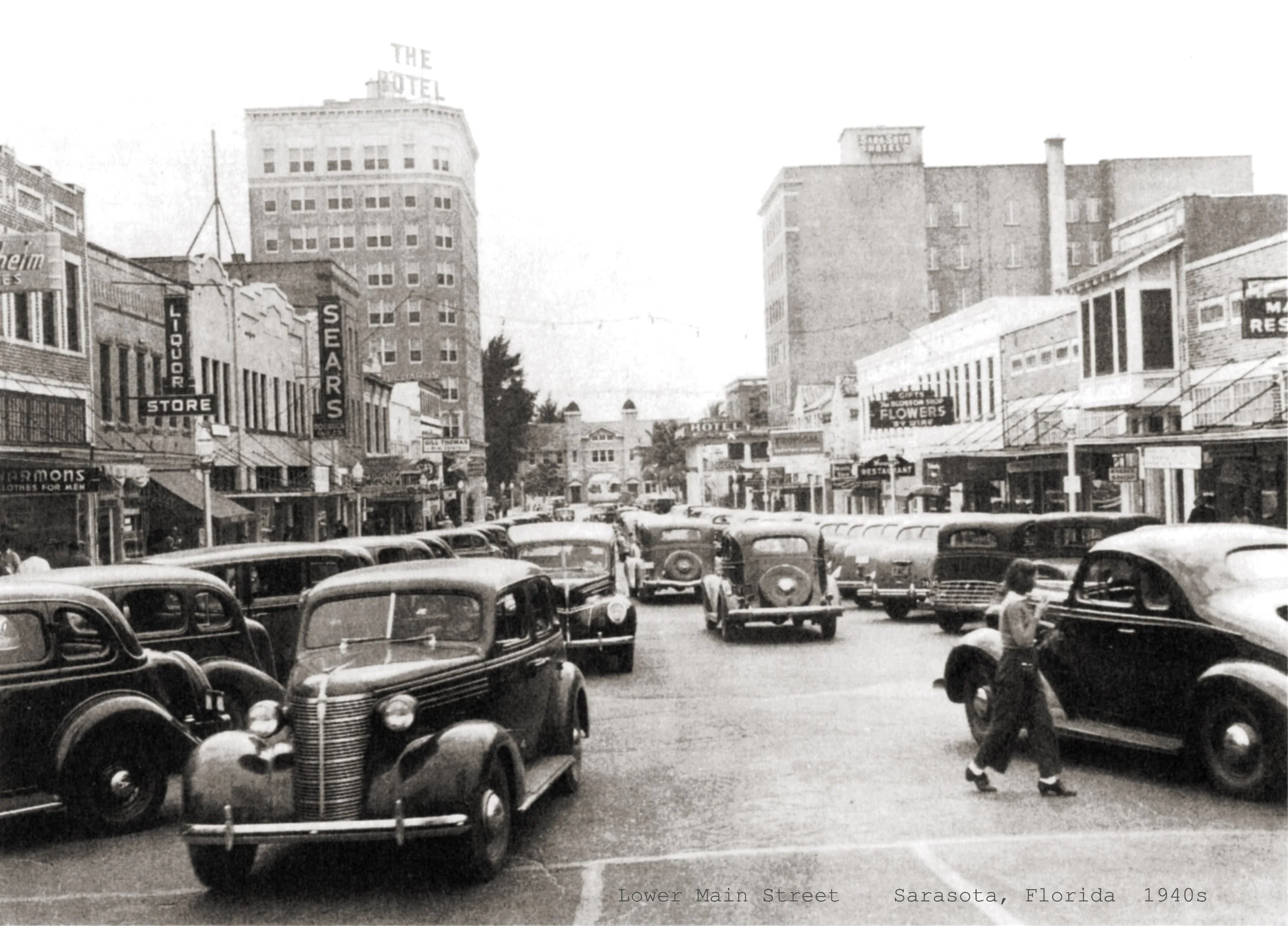 Black and white photo of Lower Main Street in Sarasota, Florida, taken in the 1940s. Vintage cars are parked along the street with some driving, and pedestrians are walking on the sidewalk. Buildings with signs for stores, a hotel, and floral shop line the street.