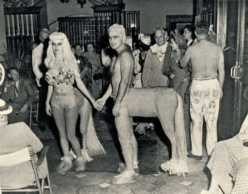 A vintage black-and-white photo of a woman with long, platinum blonde hair and a man with platinum blonde hair holding hands inside a restaurant or banquet hall. The woman is wearing tight jeans and a floral top, while the man is shirtless, with a surreal creature with the body of a horse and human arms attached behind him. There are several people seated and standing in the background, dressed in vintage attire.