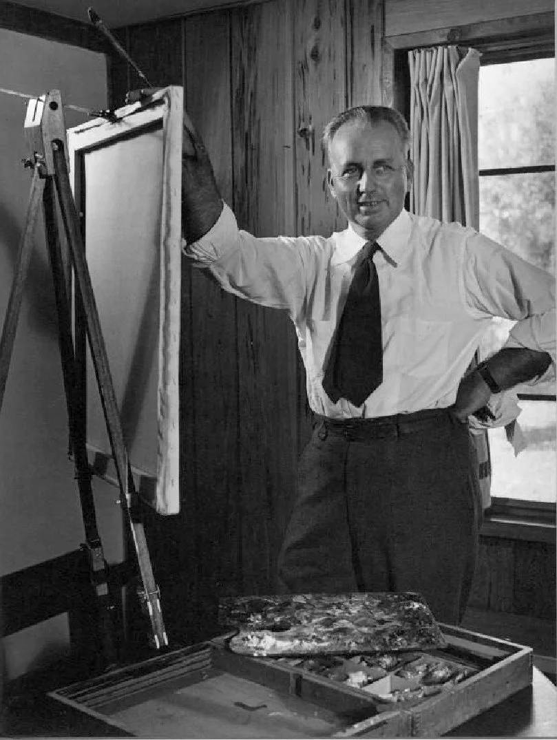 A man in a white shirt and tie stands in a room with wood-paneled walls, holding a paintbrush and standing next to an easel with a blank canvas.