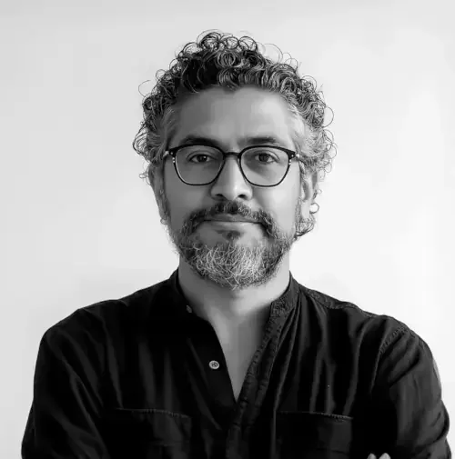 Un portrait d'un homme avec des cheveux bouclés, une barbe et des lunettes, portant une chemise noire, en noir et blanc.