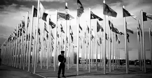 Un homme en uniforme se tient devant une rangée de nombreux drapeaux de différents pays, tous en bannière, agités par le vent, sous un ciel nuageux.