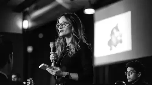 Une femme avec des lunettes parle lors d'une présentation ou d'une conférence, tenant un microphone et un carnet, en noir et blanc.