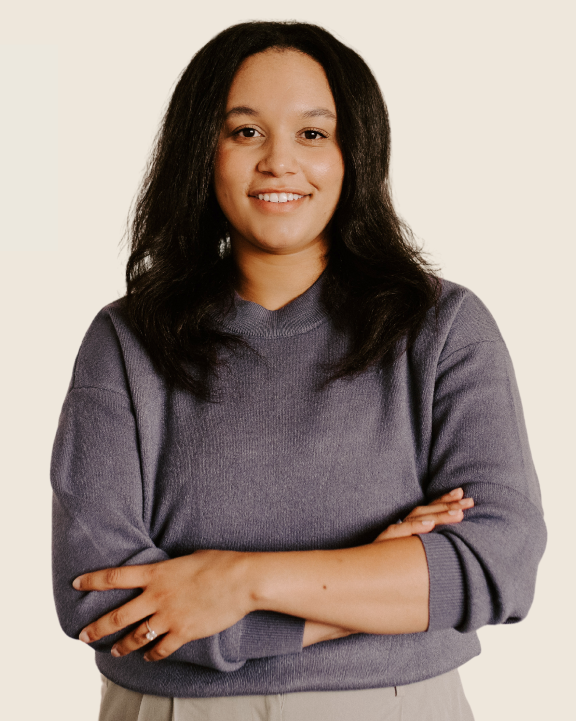 A woman with shoulder-length black hair, smiling and wearing a purple sweatshirt with arms crossed, standing against a plain off-white background.