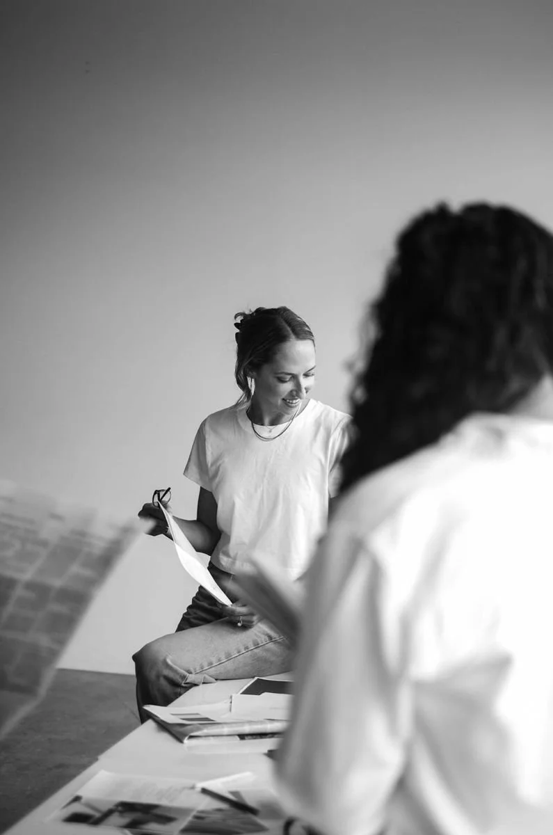 Black and white photo of a woman sitting and holding papers, smiling and looking at another person whose back is turned, in an office or workspace setting with scattered documents on a table.