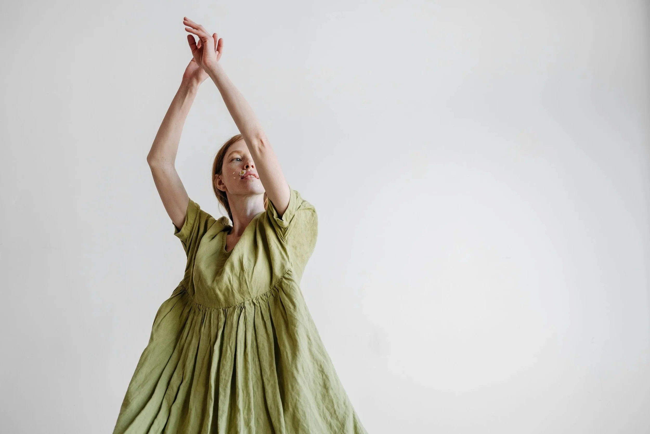 Woman in a loose, green dress raising her arms and crossing her wrists, standing against a plain white background.