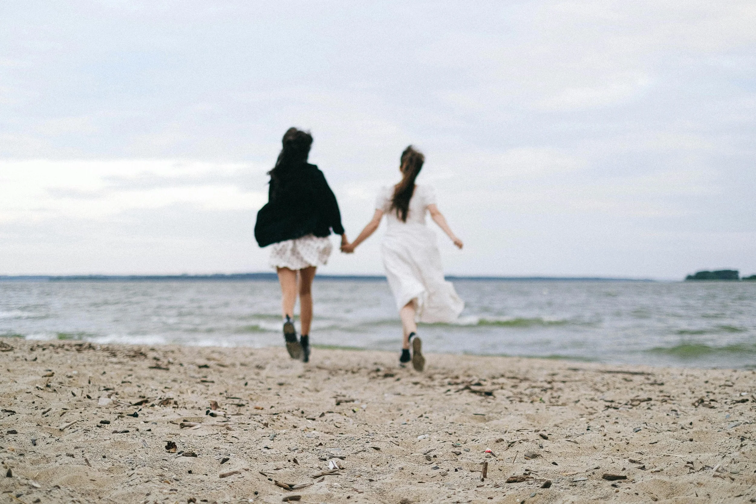 Two women holding hands while running along a beach shore during cloudy weather.