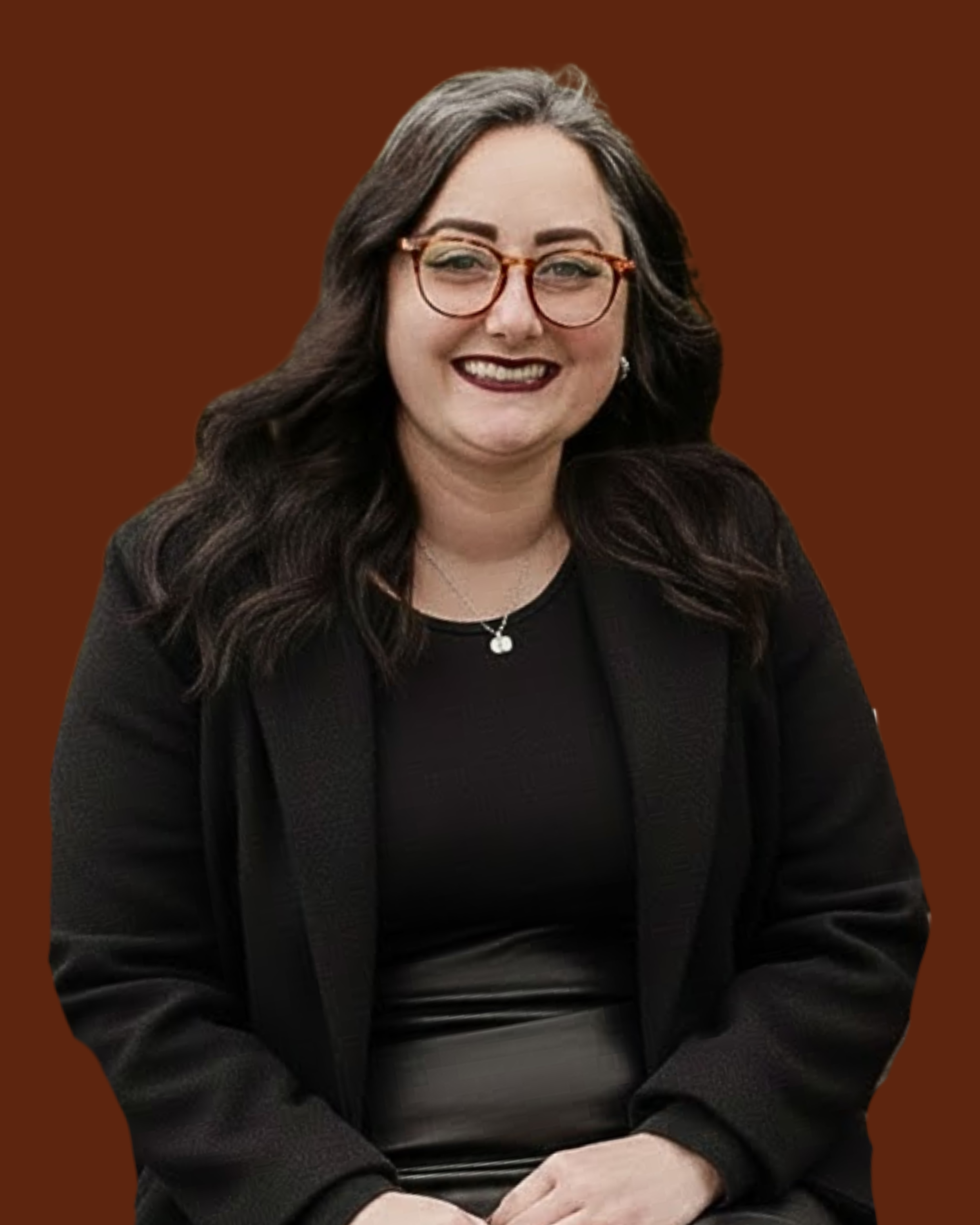 A woman with dark wavy hair, glasses, and a black outfit, sitting and smiling against a brown background.