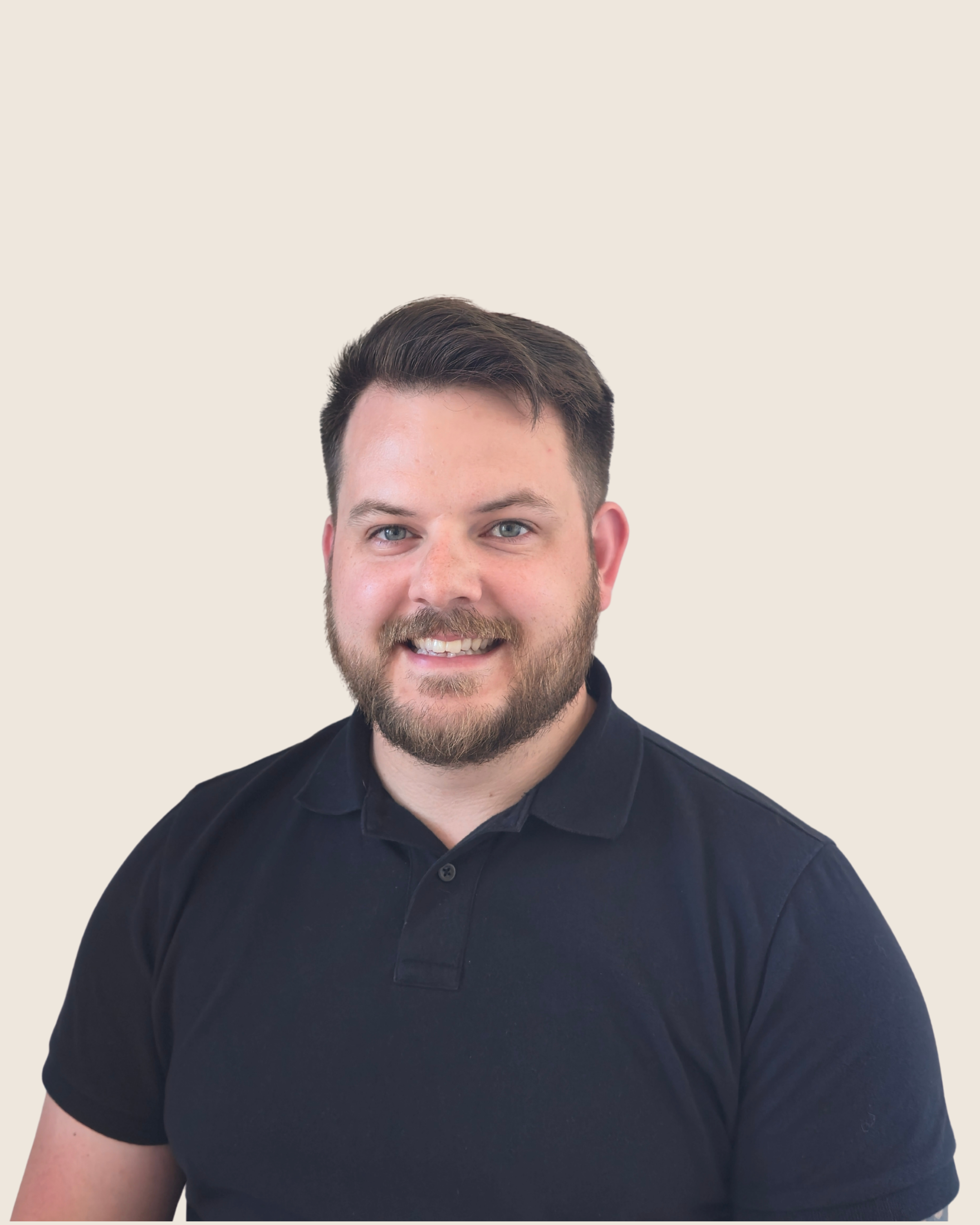Portrait of a smiling man with short dark hair, beard, wearing a black polo shirt against a plain light-colored background.