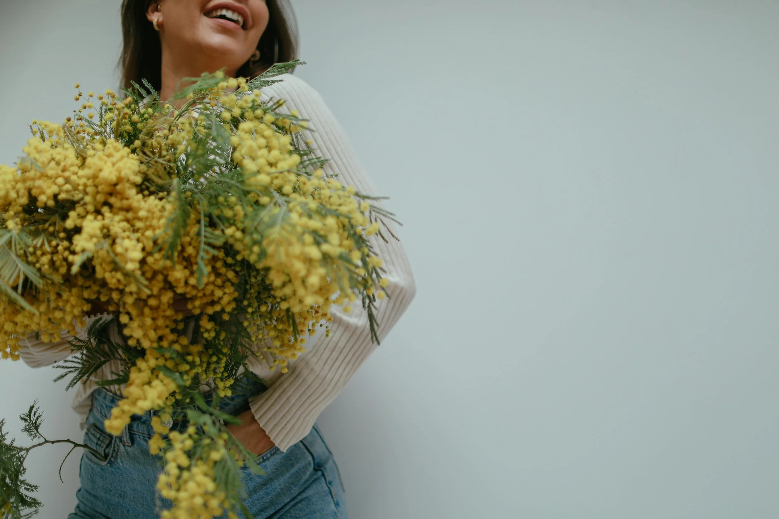 Woman smiling while holding a large bouquet of yellow flowers against a plain light-colored wall.