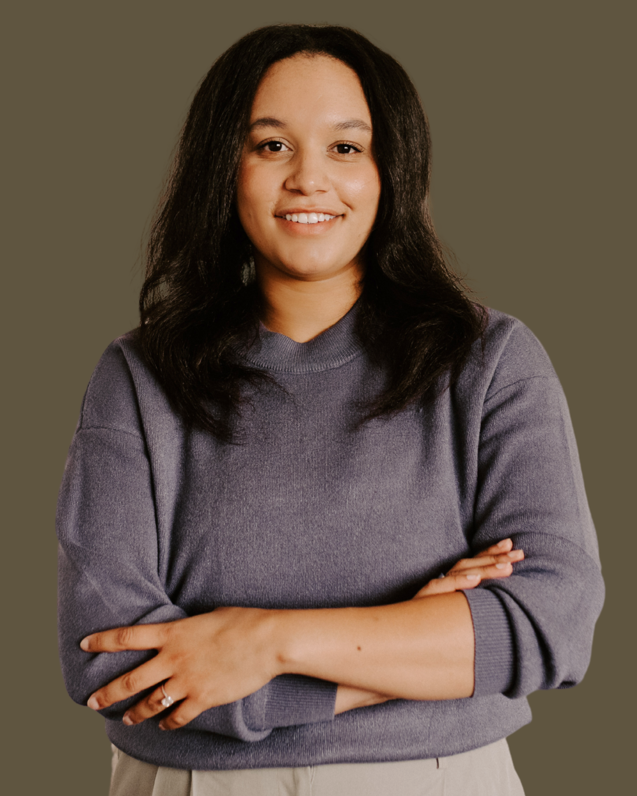 A young woman with shoulder-length dark hair wearing a gray sweater and beige pants, standing with her arms crossed and smiling at the camera.