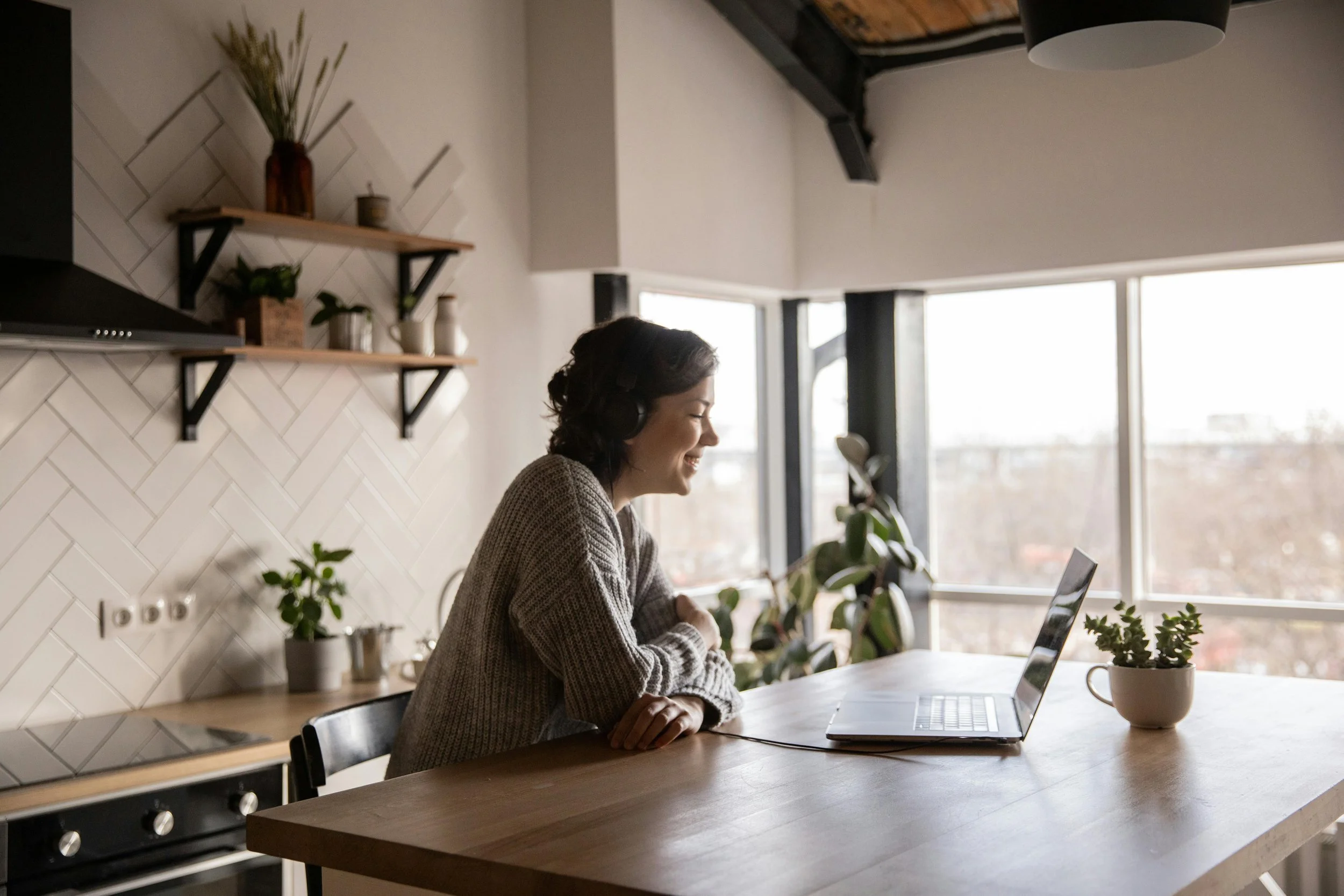 A woman is smiling and wearing headphones while looking at her laptop in a bright kitchen or dining area with large windows and potted plants.