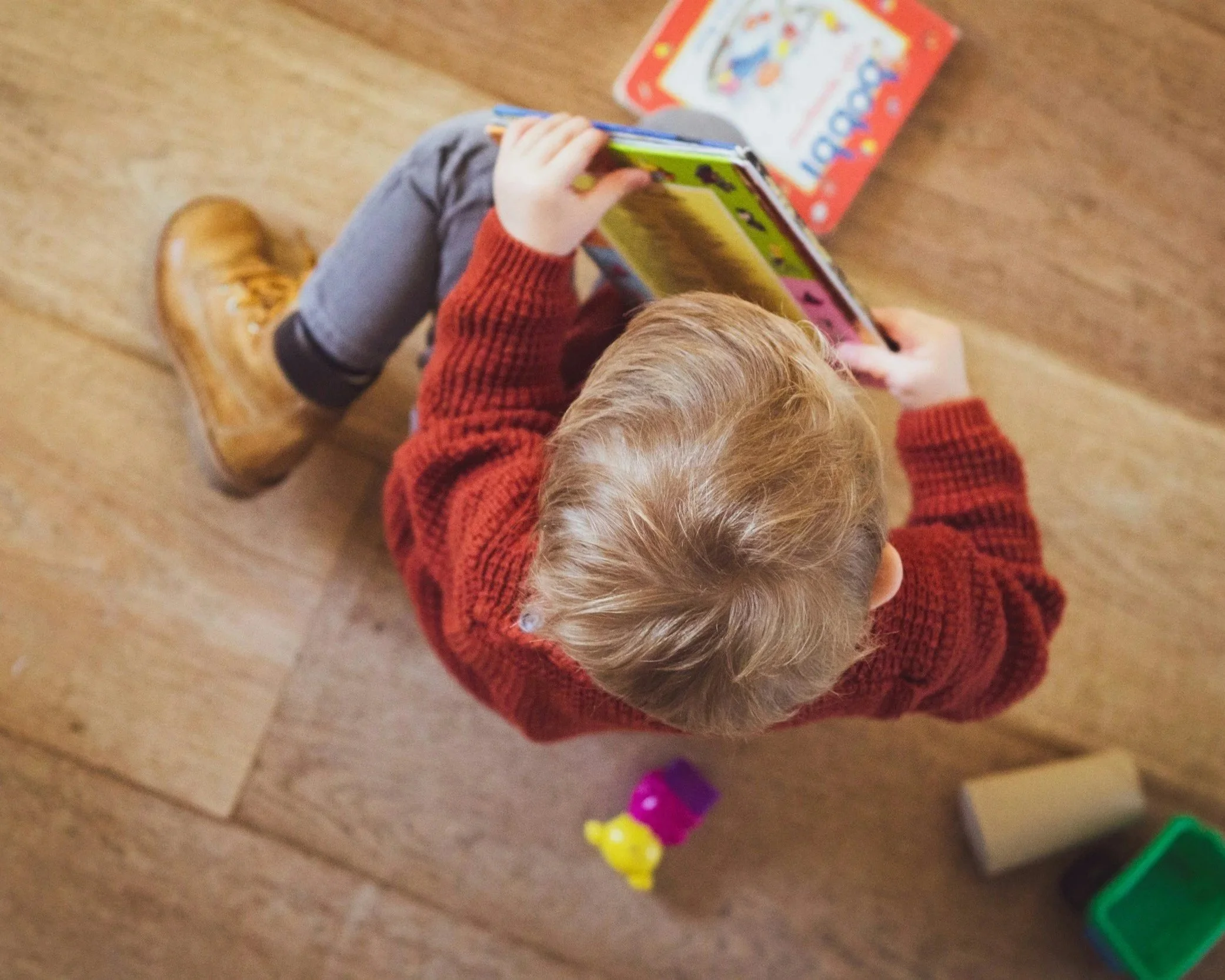 Child in red sweater playing with colorful building blocks on a wooden floor.