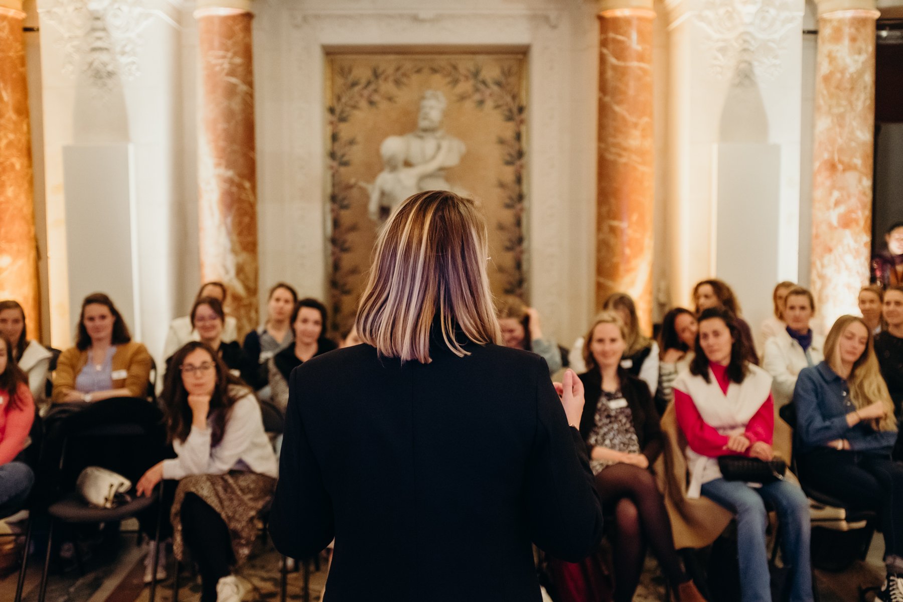 Femme donnant une présentation à un groupe dans une salle ornée de colonnes en marbre et d'un mur décoratif avec une sculpture antique.