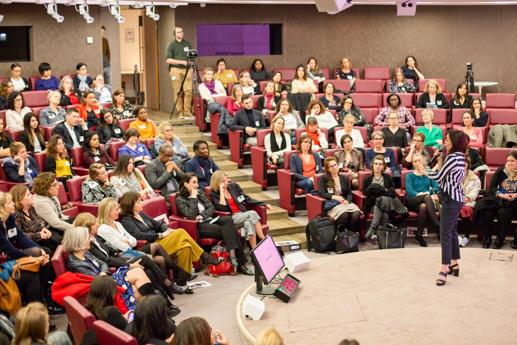 Une femme parle dans un micro devant un public au sein d'une salle de conférence avec des sièges rouges et un écran.