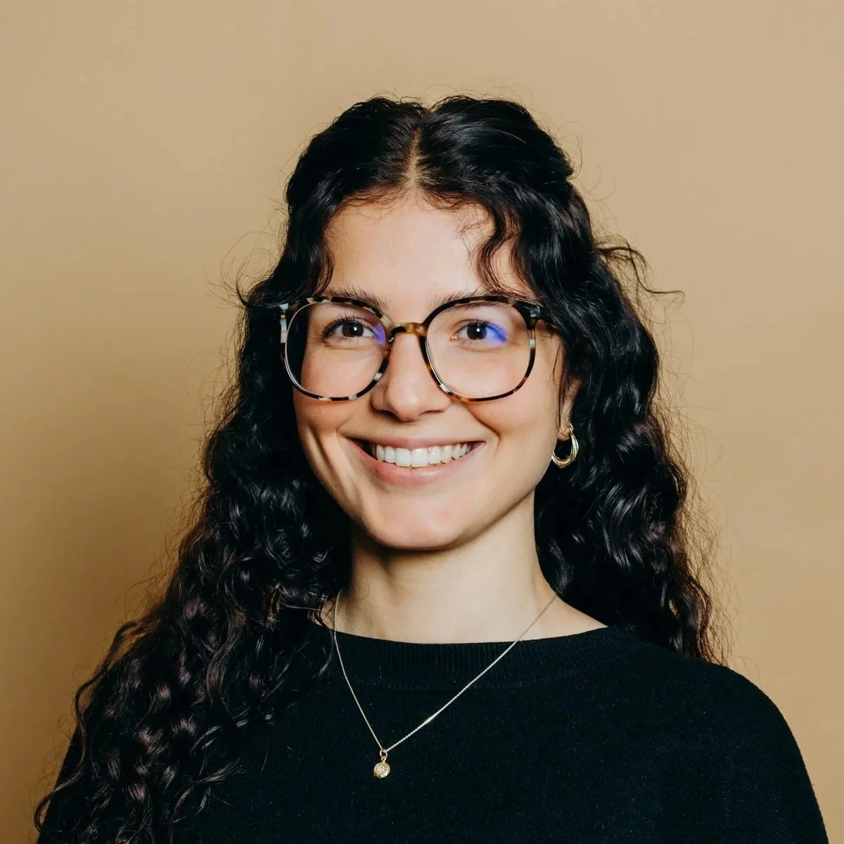 Jeune femme souriante portant des lunettes et un col noir, avec des boucles de cheveux longs et foncés, portant des boucles d'oreilles en or et une chaîne avec un pendentif, devant un fond beige.