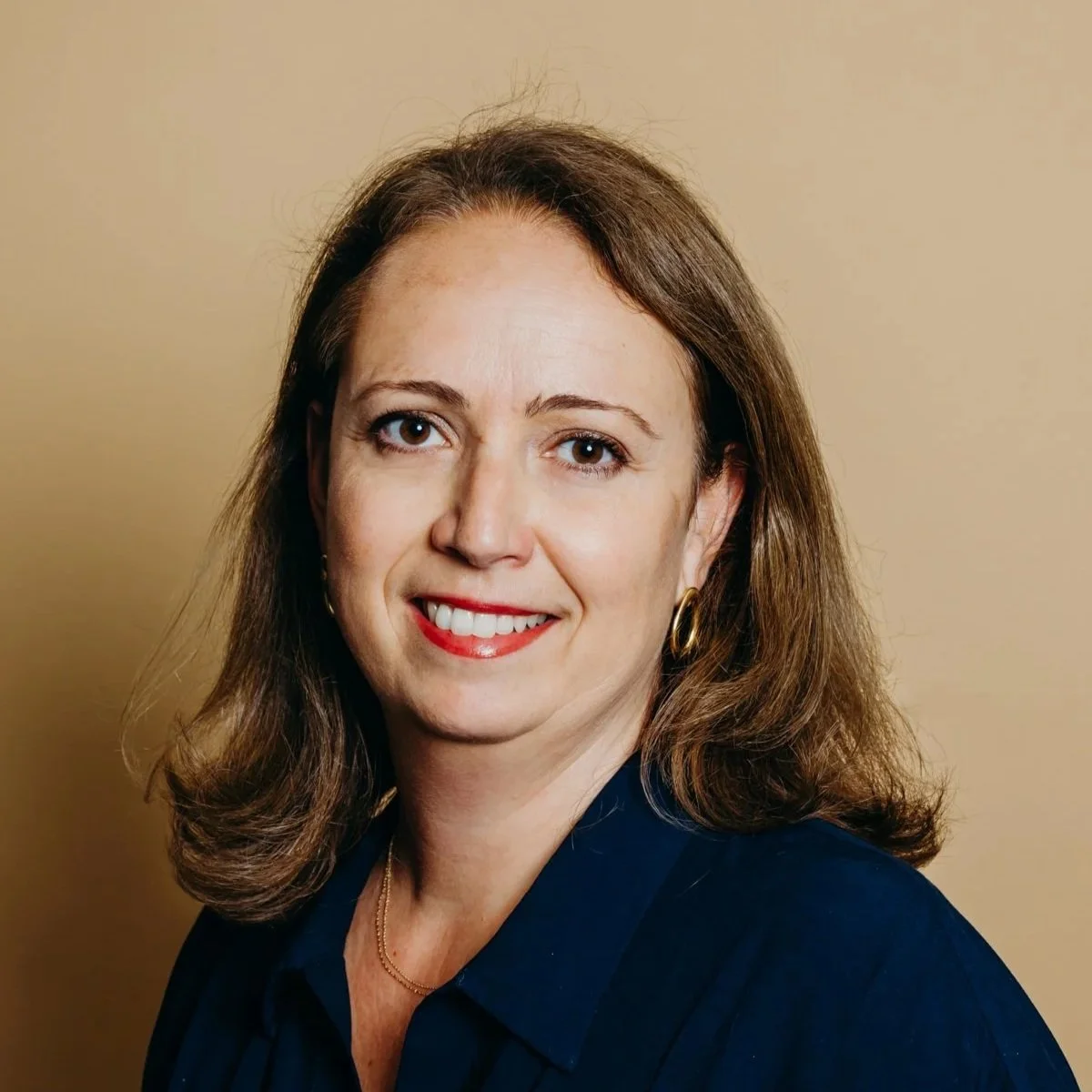 Portrait d'une femme aux cheveux bruns, souriante, portant une chemise bleue et des boucles d'oreilles en or, avec un fond beige.