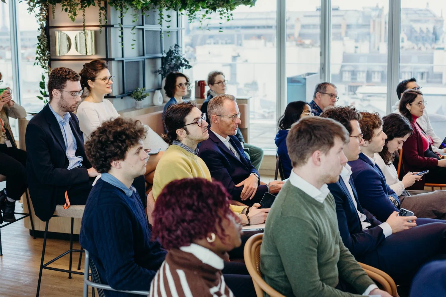 Un groupe de personnes assises lors d'une conférence ou réunion dans une salle moderne avec de grandes fenêtres laissant entrer la lumière naturelle, et des plantes comme décoration interne.