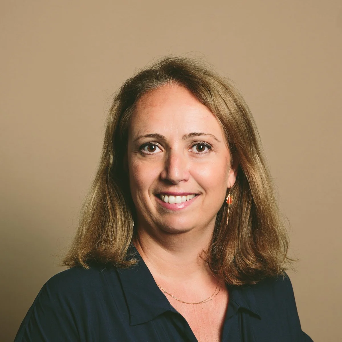 Portrait d'une femme souriante avec des cheveux bruns, portant un haut noir et des boucles d'oreilles, contre un fond beige.