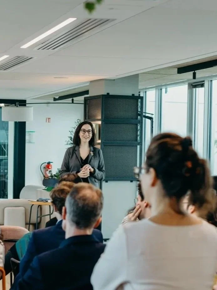 Une femme souriante parle lors d'une conférence ou d'une présentation devant un groupe de personnes dans une salle moderne avec de grandes fenêtres et un éclairage naturel.