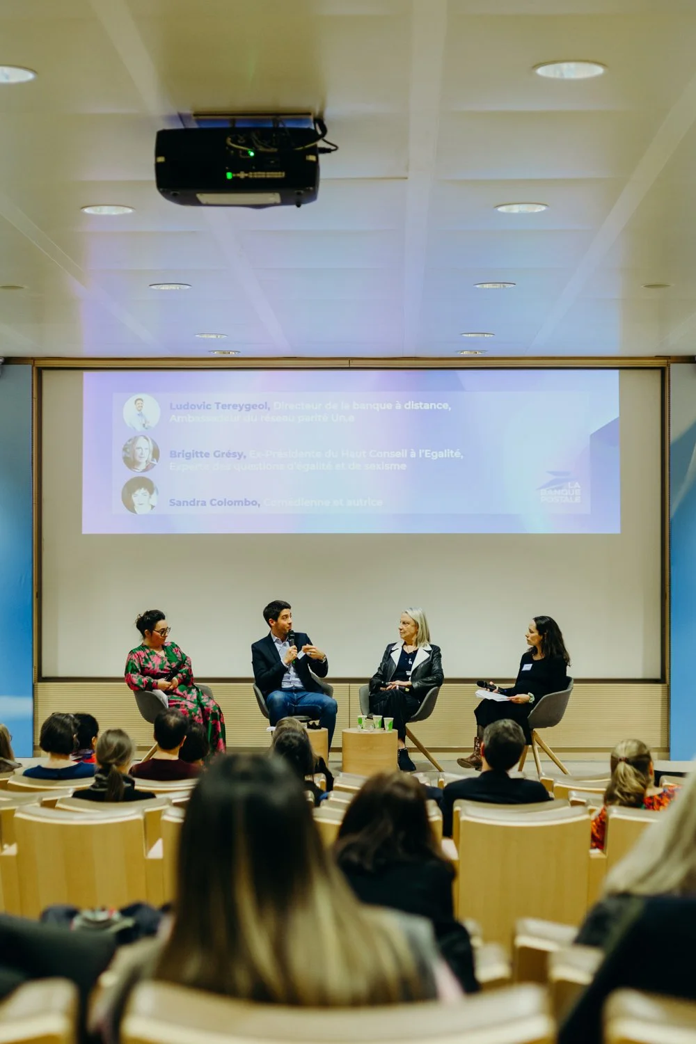 Une table ronde avec quatre femmes en plein débat lors d'une conférence, avec un public en arrière-plan et un grand écran derrière elles affichant des noms et des titres.