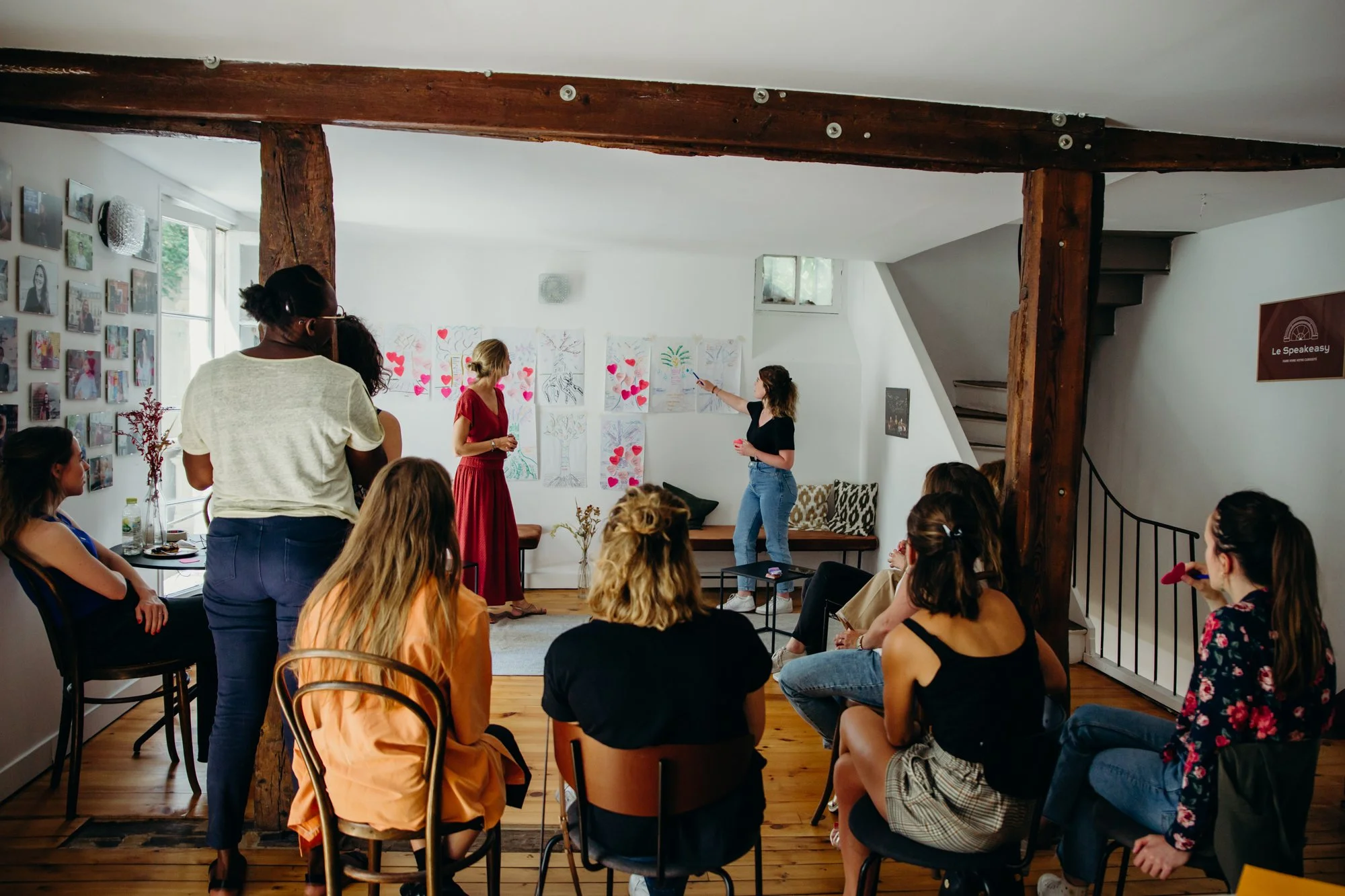 Groupe de personnes assises et debout dans une salle accueillante lors d'une présentation ou d'un atelier, avec des œuvres d'art sur les murs et une ambiance conviviale.