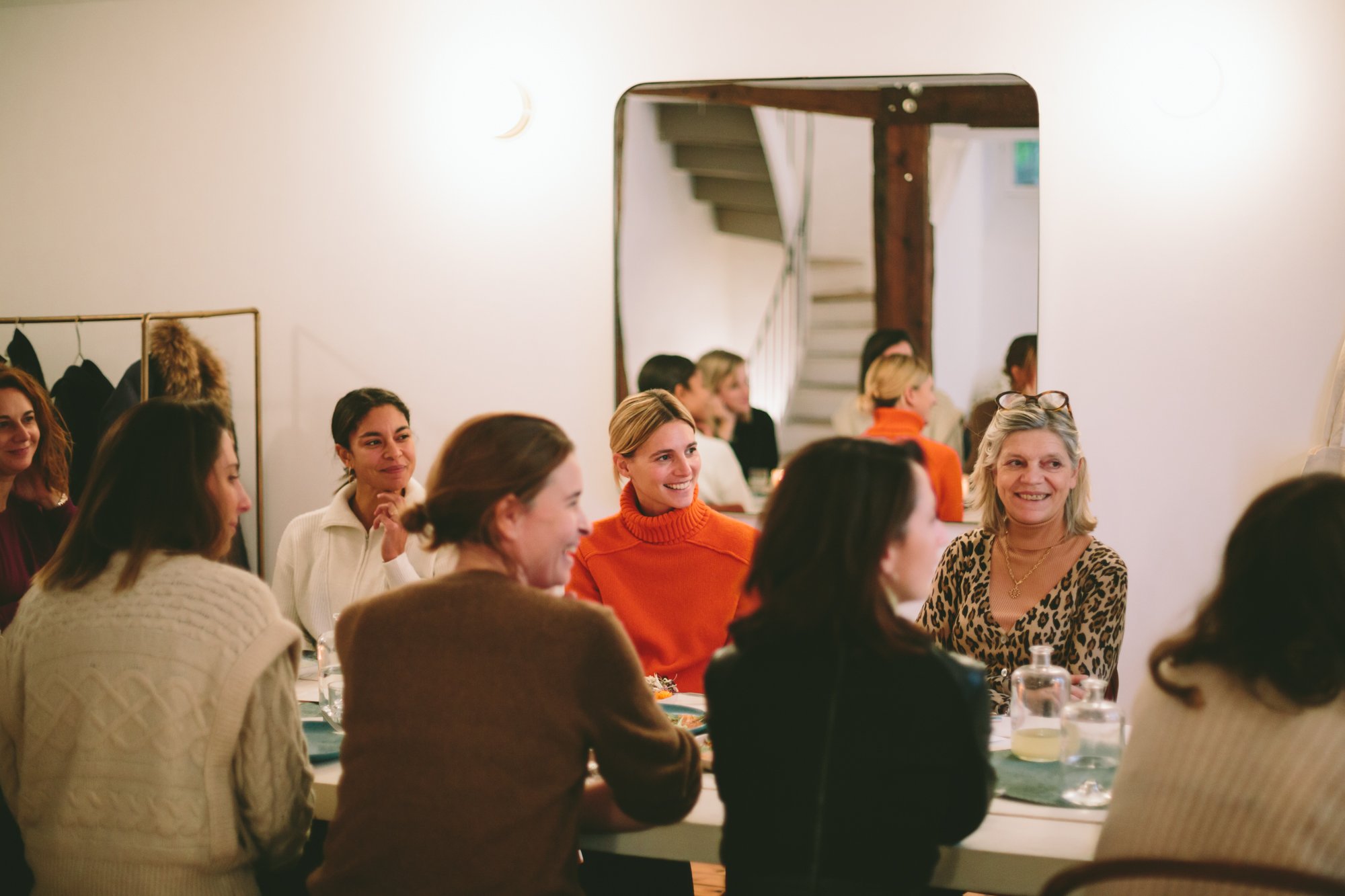 Groupe de femmes souriantes autour d'une table lors d'une rencontre ou d'un repas convivial à l'intérieur, avec un miroir laissant apparaître un escalier en arrière-plan.