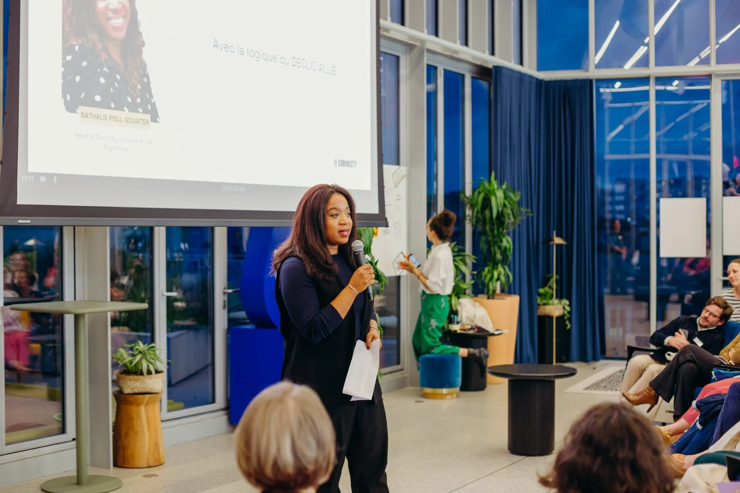 Une femme parle lors d'une présentation dans une salle moderne avec des grandes fenêtres, des plantes et des personnes assises dans l'audience.