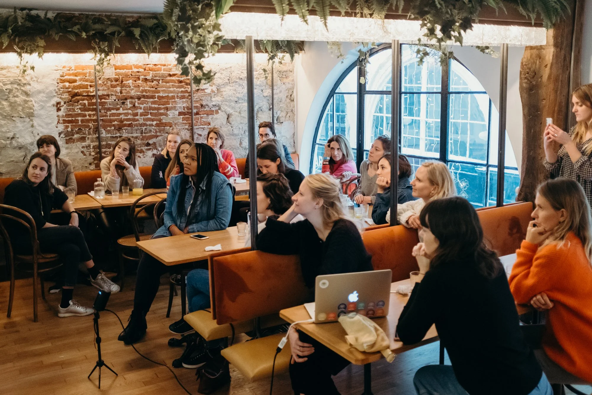 Un groupe de jeunes femmes assises dans un café ou un espace de coworking, certaines écoutant attentivement, d'autres utilisant un ordinateur portable ou prenant des photos, avec un décor intérieur en briques et un grand mur en verre