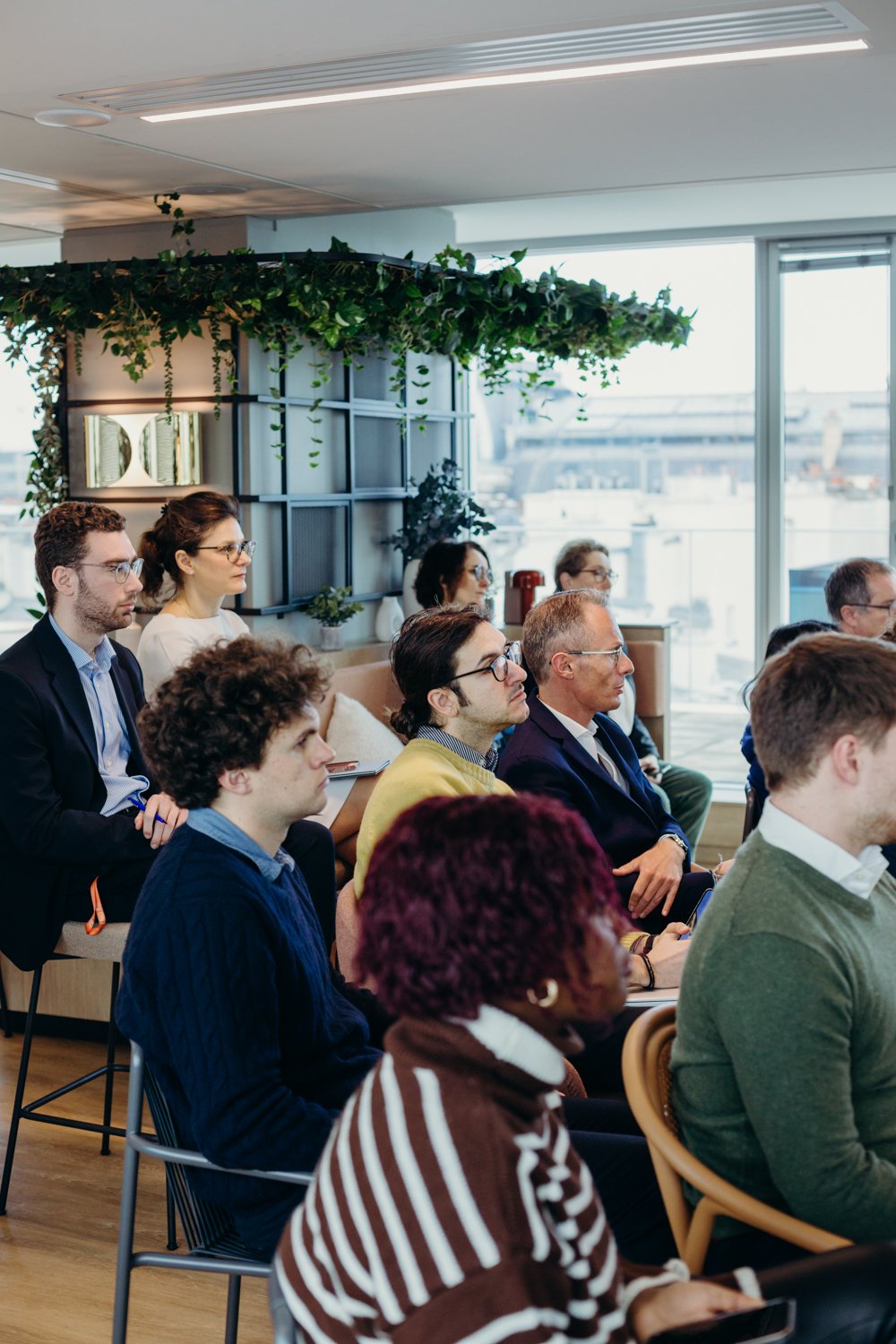 Groupe de personnes assises lors d'une réunion ou conférence dans une salle moderne avec de grandes fenêtres et décor végétal.