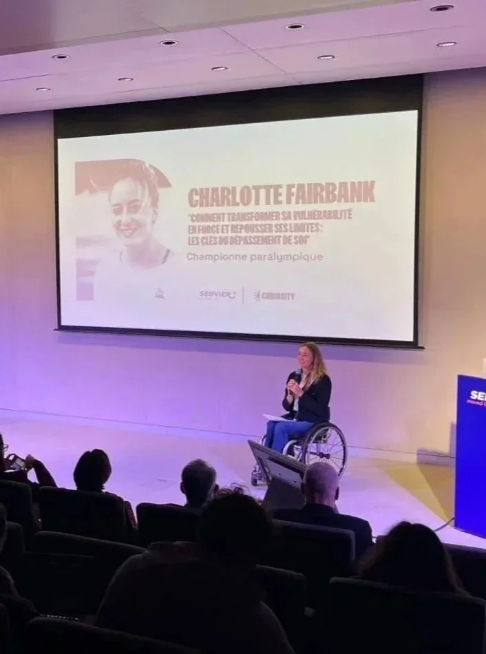 Une femme en fauteuil roulant parlant lors d'une conférence sur l'inclusion des personnes en situation de handicap, avec un grand écran derrière elle affichant un slide à propos de Charlotte Fairbank.