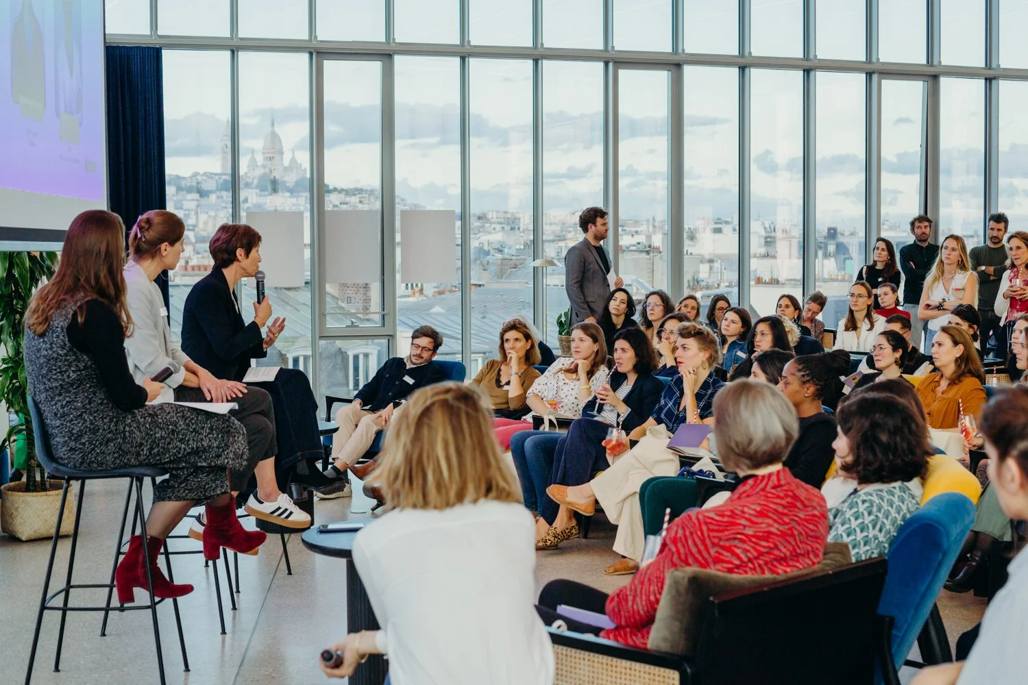 Une conférence ou un panel avec plusieurs intervenants et un public diversifié dans une salle moderne avec de grandes fenêtres offrant une vue sur la ville de Paris.