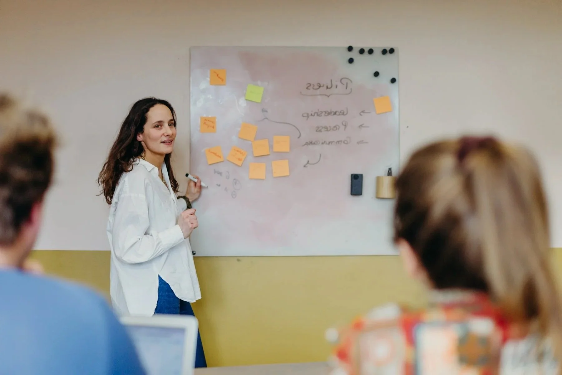 Une femme enseigne devant un tableau blanc avec des Post-it en classe, des personnes assises à une table l'écoutent.