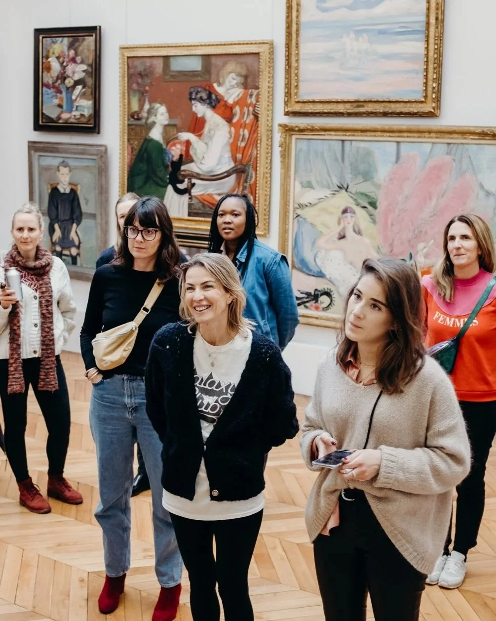 Un groupe de personnes, principalement des femmes, dans une galerie d'art, regardant et discutant autour de peintures accrochées aux murs.