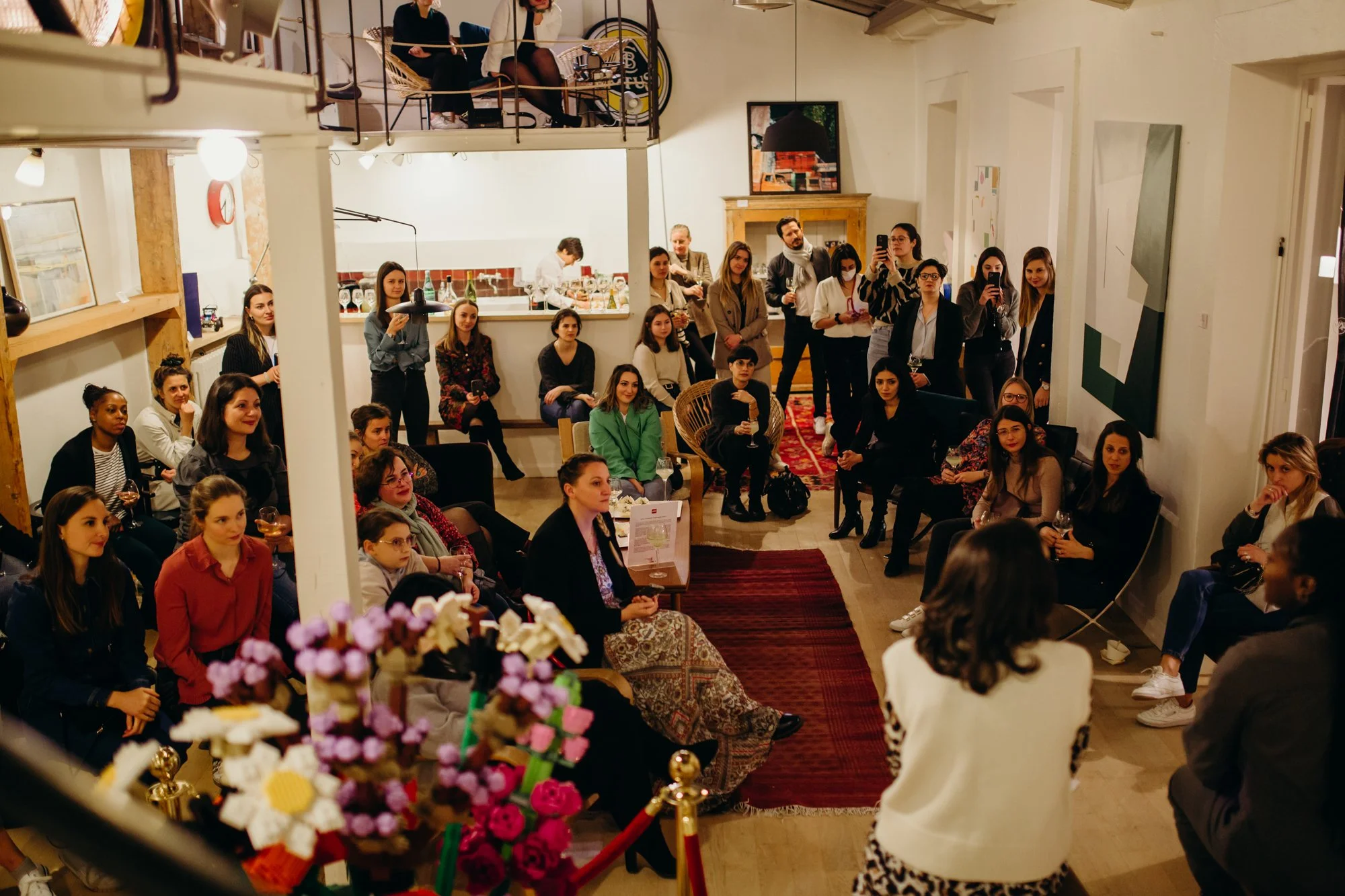 Une salle remplie de personnes assises et debout, probablement lors d'une présentation ou d'un événement social, avec un décor intérieur chaleureux et des œuvres d'art accrochées aux murs.