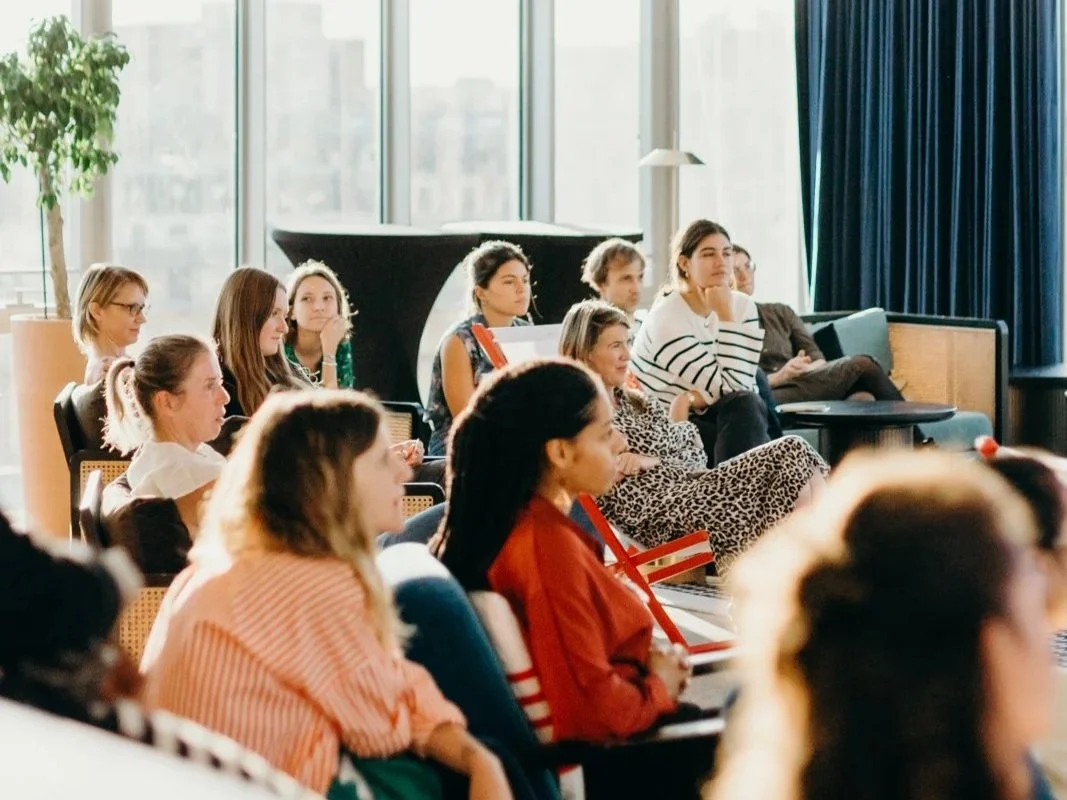 Groupe de personnes assises dans une salle de conférence ou un auditorium, écoutant une présentation ou un discours, avec de grandes fenêtres en arrière-plan.