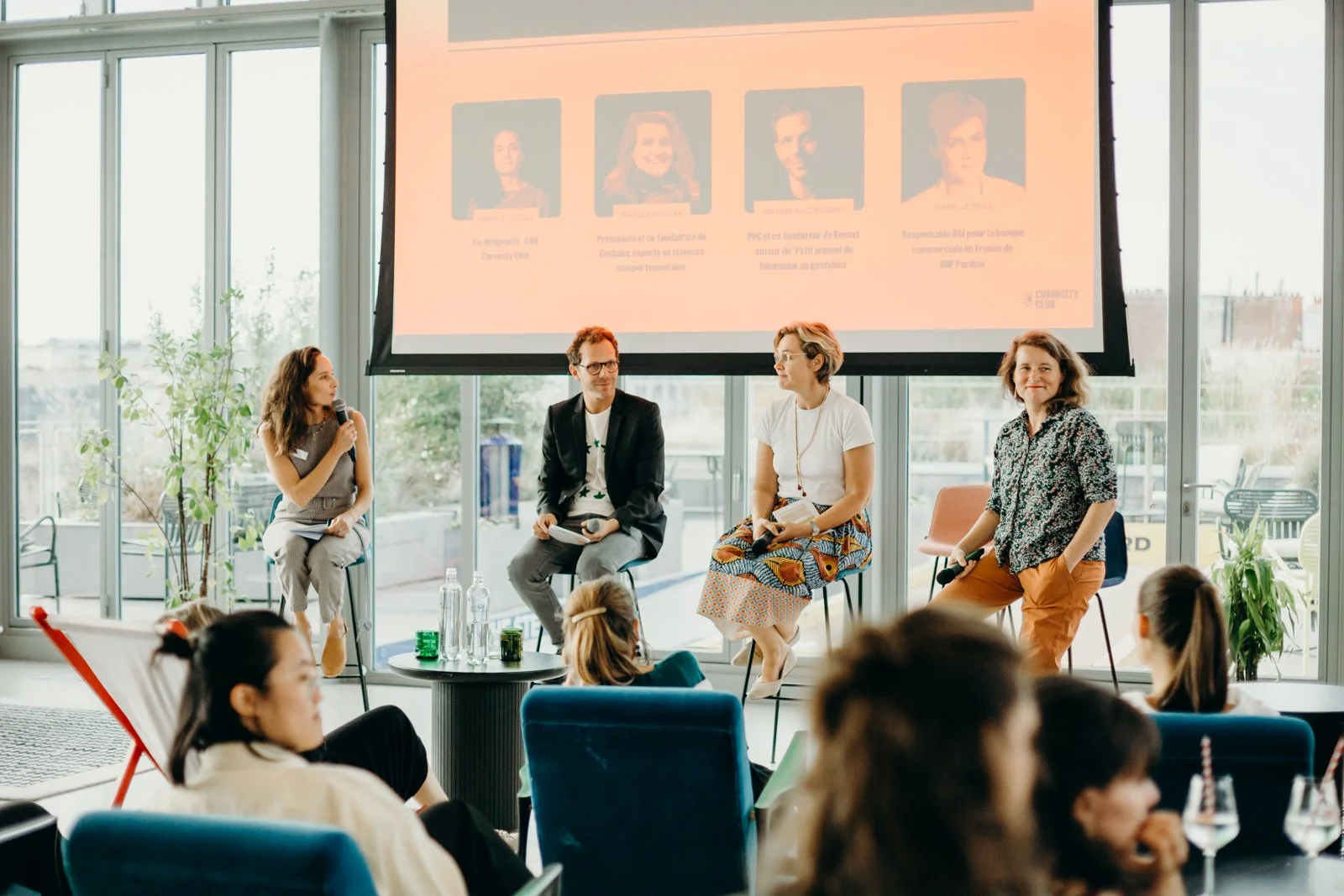 Une table ronde avec quatre personnes qui parlent lors d'une conférence ou panel, avec un public qui écoute. Un grand écran derrière affiche des portraits et textes.
