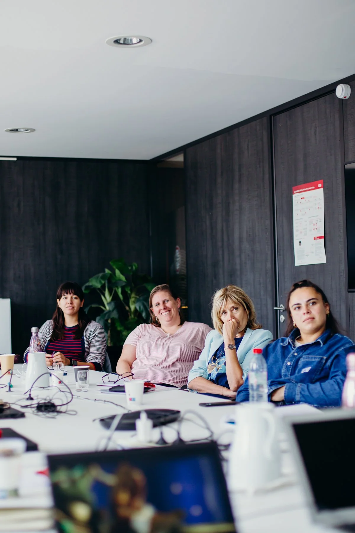 Quatre femmes assises autour d'une table lors d'une réunion, avec des ordinateurs portables, des bouteilles d'eau, des tasses en papier, dans une salle de conférence moderne.