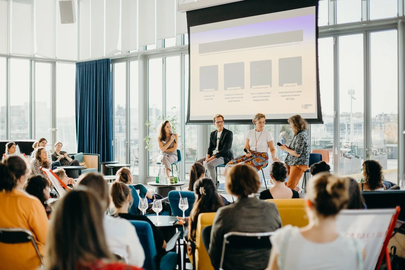 Une conférence ou panel avec quatre personnes en discussion, assises face à une audience dans une salle lumineuse et moderne, avec un grand écran derrière eux et de larges fenêtres offrant une vue sur la ville.