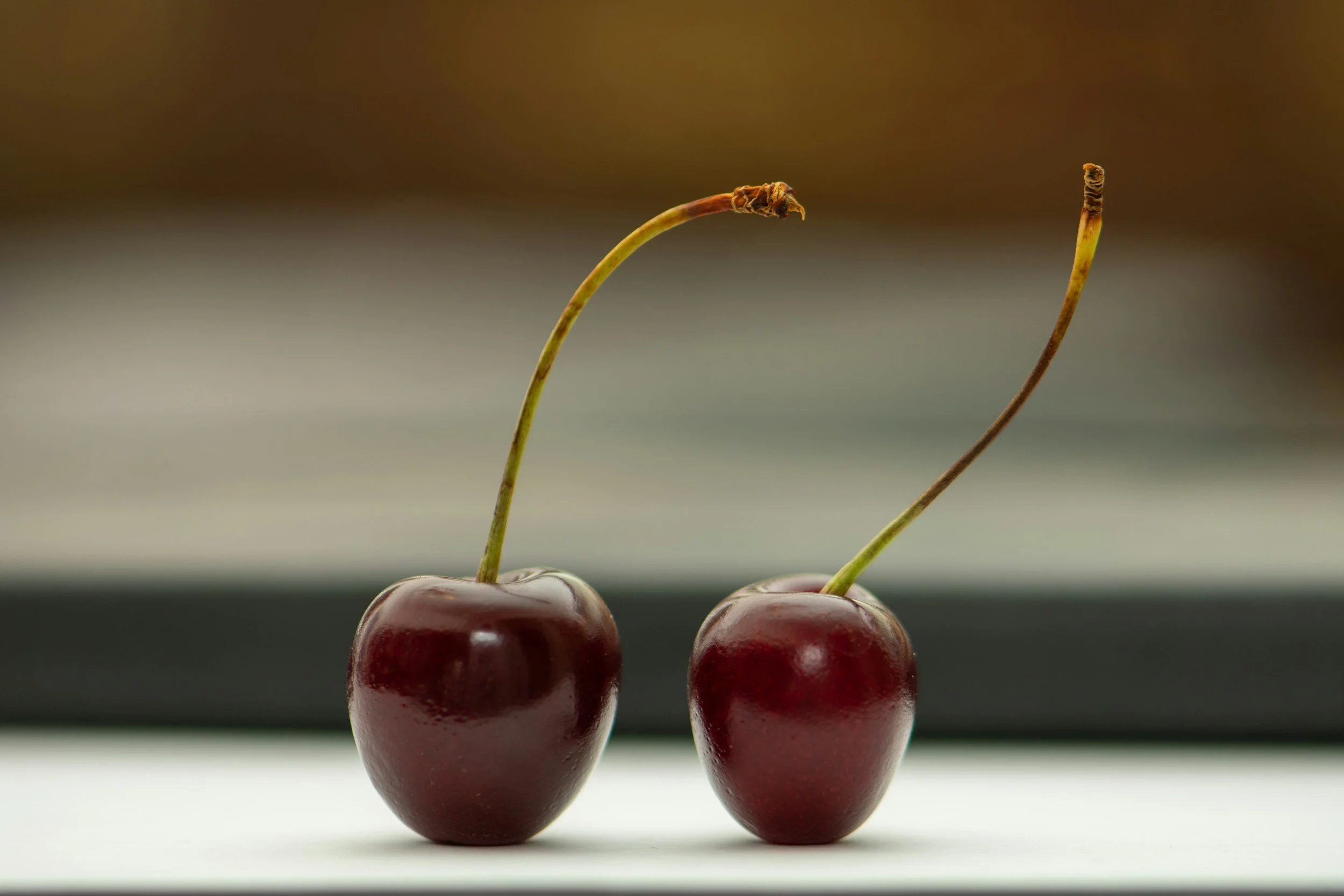 Two cherries with stems, one on the left and one on the right, resting on a flat surface against a blurred background.